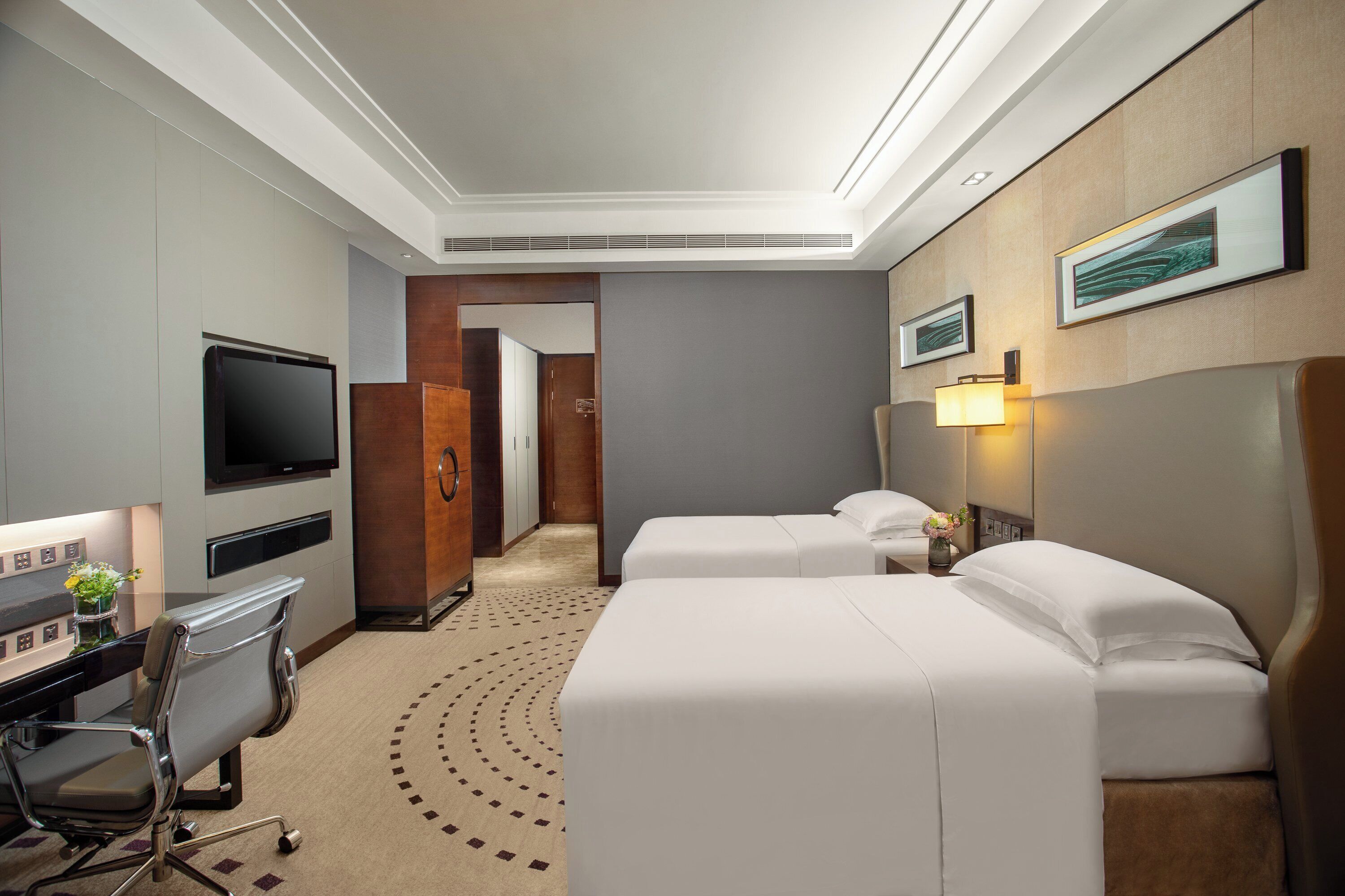 Quarto de hotel com duas camas, uma escrivaninha com cadeira, televisão e banheiro visível através de uma porta aberta.