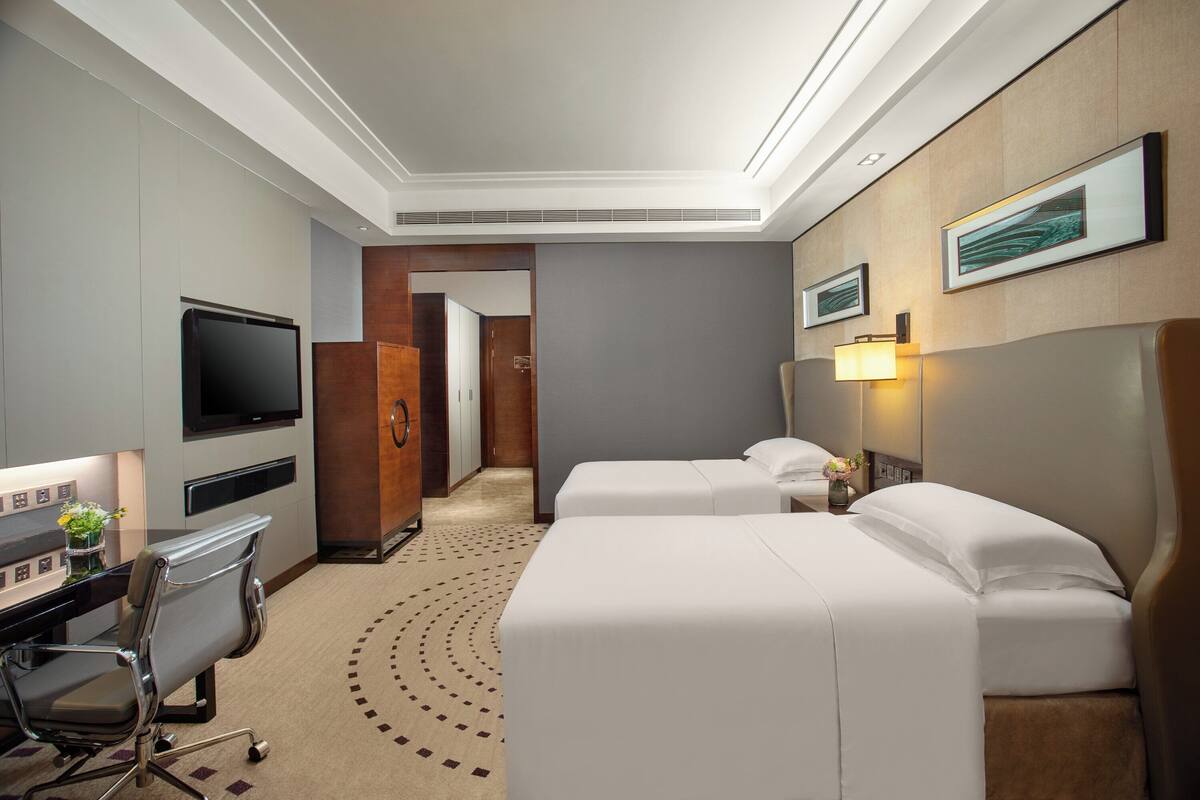 A hotel room with two beds, a desk with a chair, a TV, and a bathroom visible through an open door.