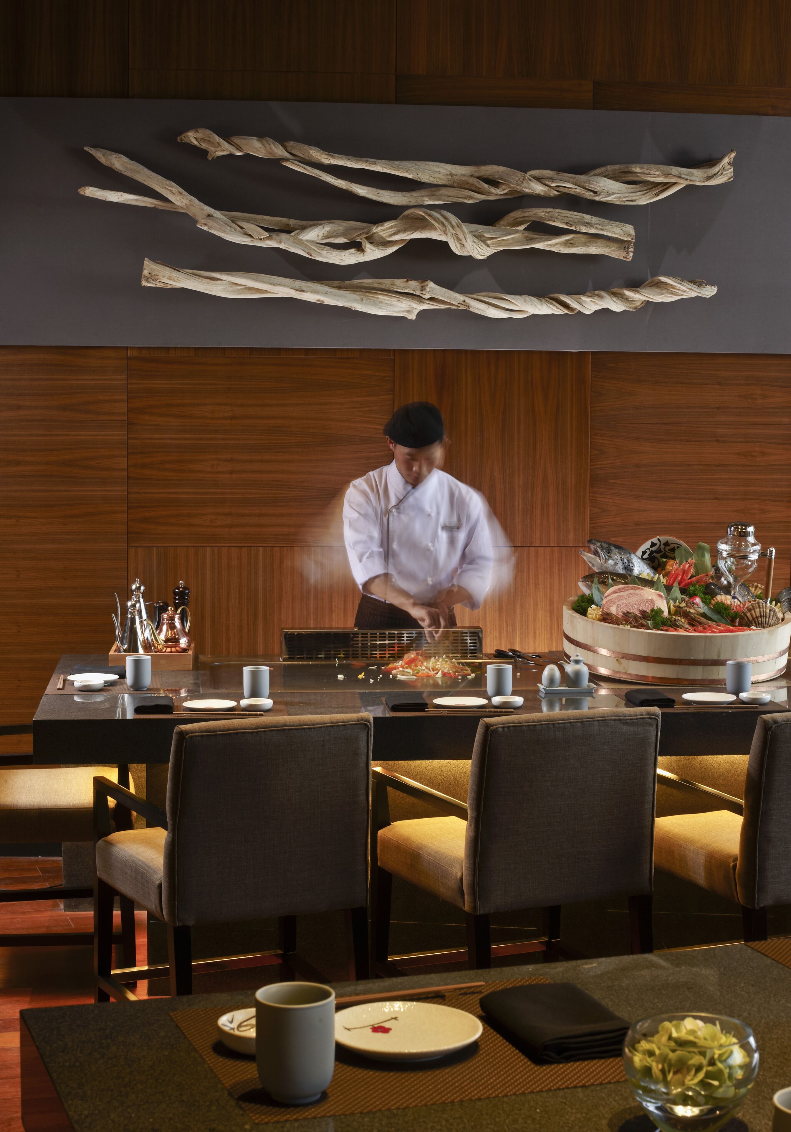 A chef preparing food at a table in a restaurant with wooden paneling and a large decorative piece above the table.
