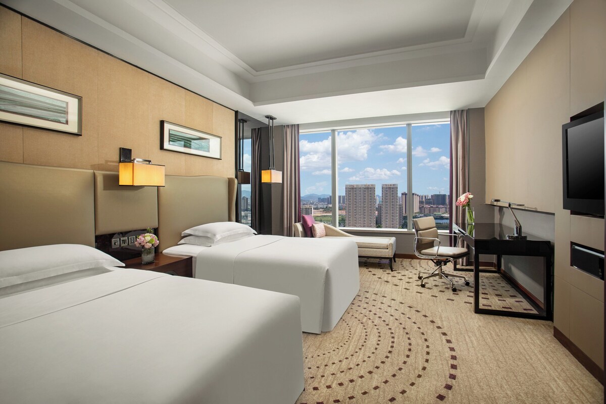 A hotel room with two beds, a desk, a chair, a TV, and a large window with a city view.
