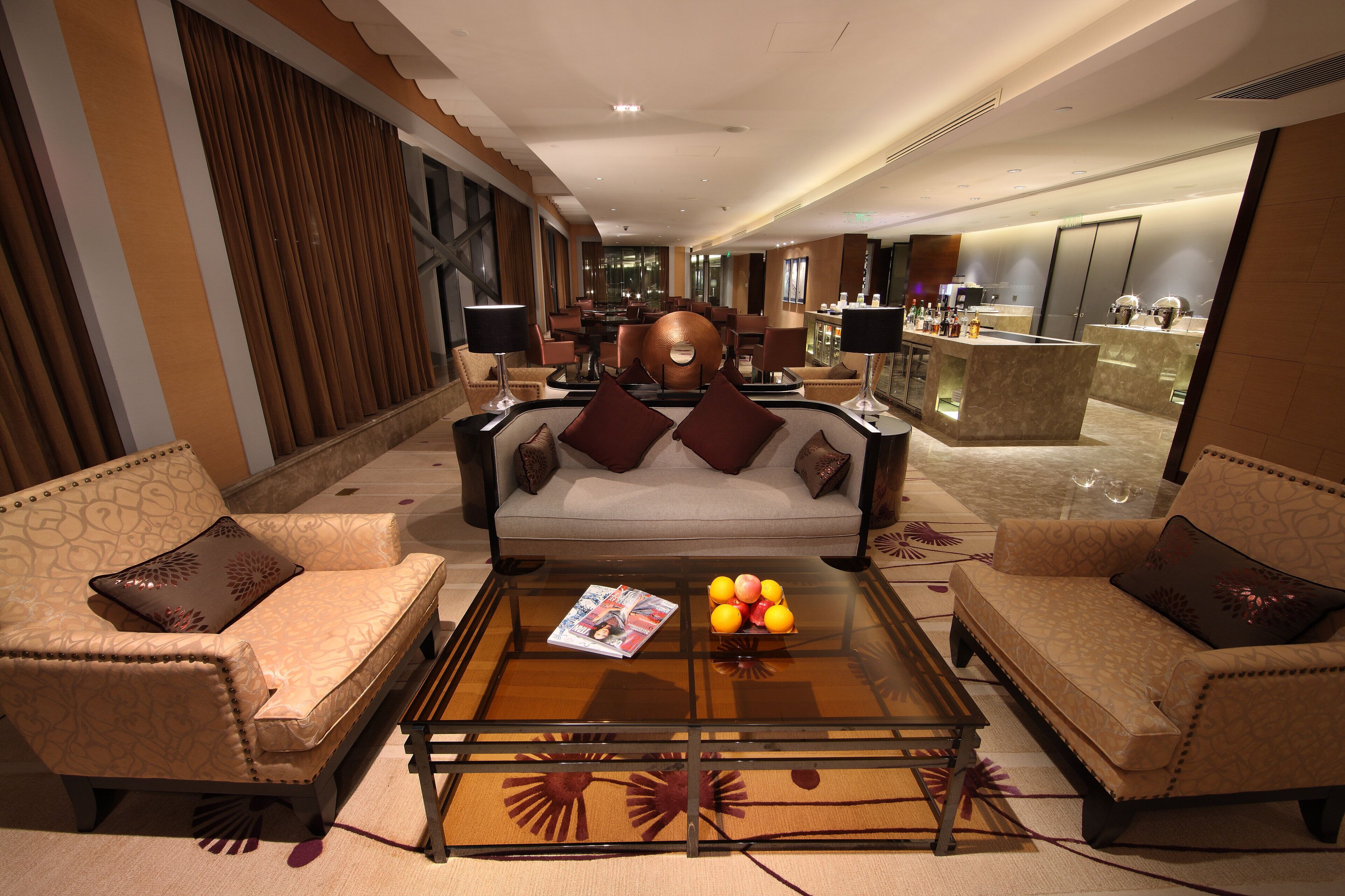 A hotel lobby with a sofa set, a coffee table with magazines, and a bar area in the background.