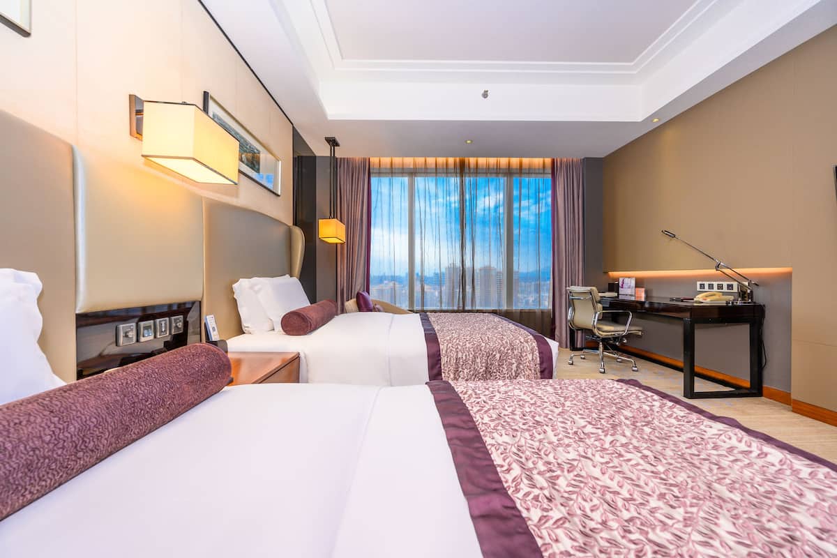 A hotel room with two beds, a desk, a chair, a lamp, and a large window with curtains.