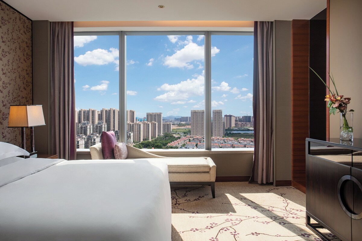 A hotel room with a large window offering a city view, a bed, a chair, and a dresser.