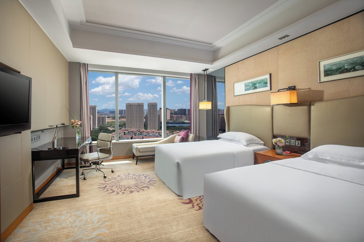 Habitación de hotel con cama, escritorio, silla, televisor y un ventanal con vista a la ciudad.