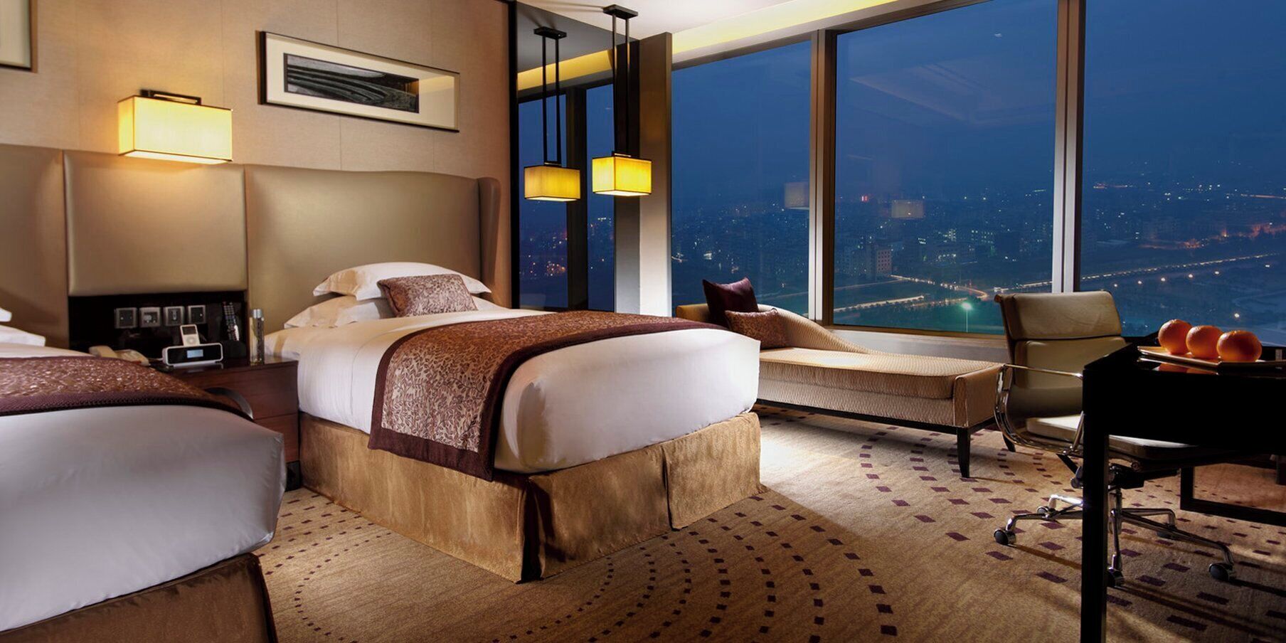 A hotel room with a large bed, a sofa, a desk with fruit, and a city view.
