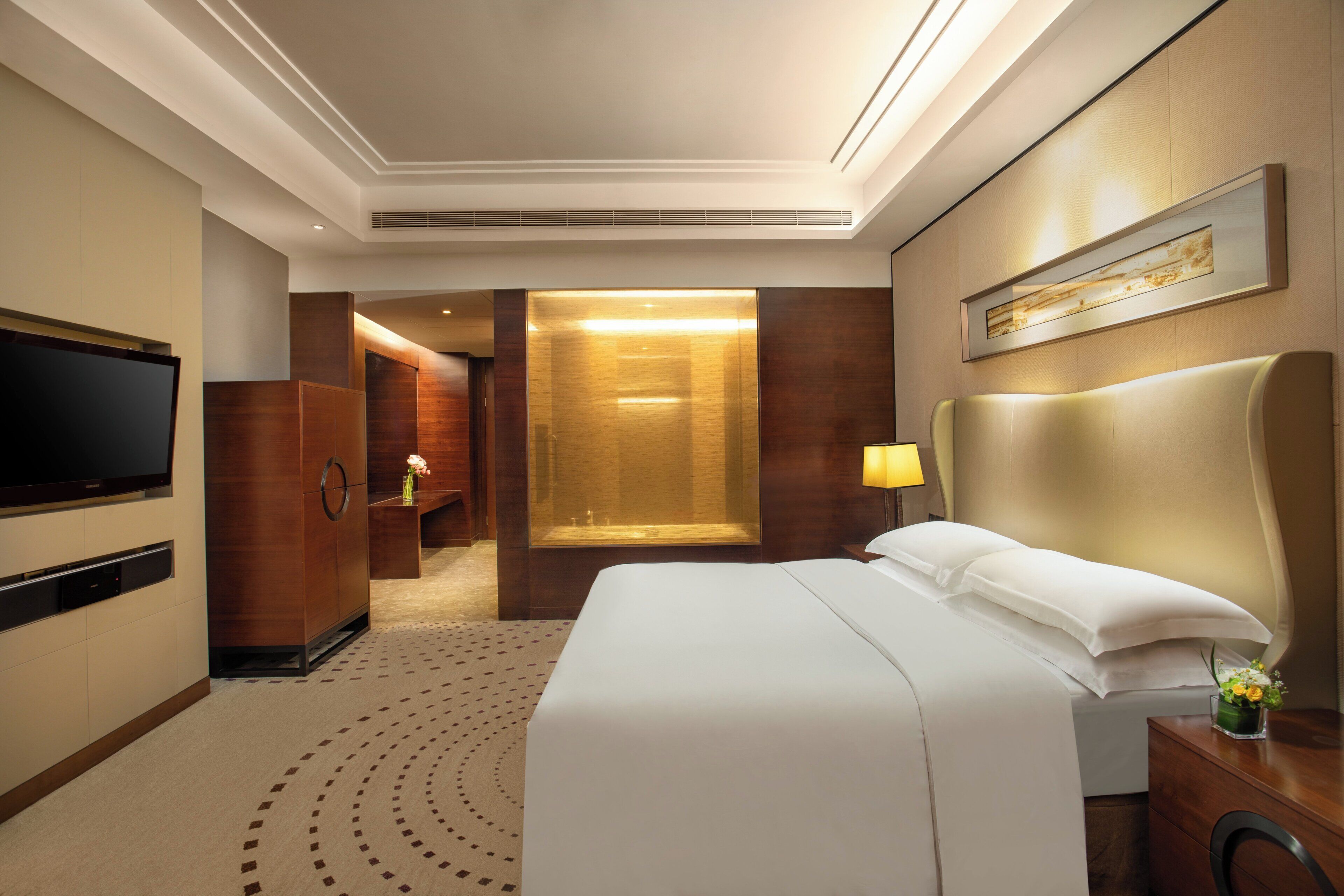 Quarto de hotel com uma cama grande, televisão, guarda-roupa de madeira, espelho e banheiro visível através de uma porta aberta.