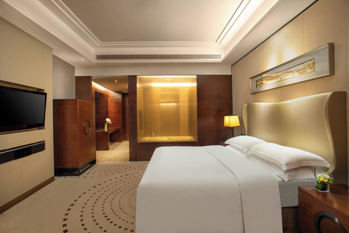 A hotel room with a large bed, a TV, a wooden wardrobe, a mirror, and a bathroom visible through an open door.