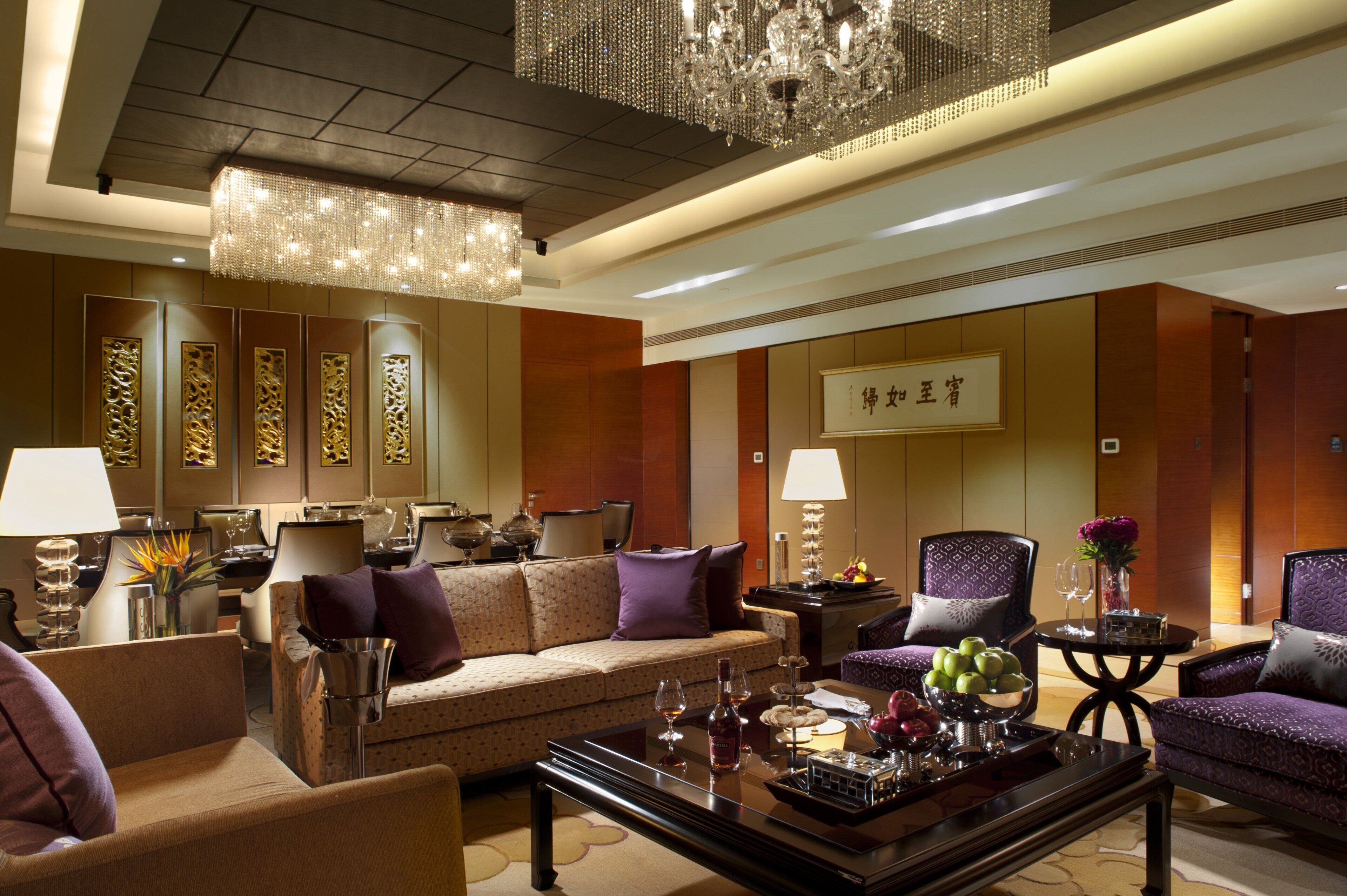 A hotel lobby with a sofa, armchairs, a coffee table with fruit and a bottle, a chandelier, and a sign with Chinese characters.