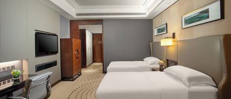 A hotel room with a bed, bedside table, lamp, TV, desk, chair, and a view of the hallway.