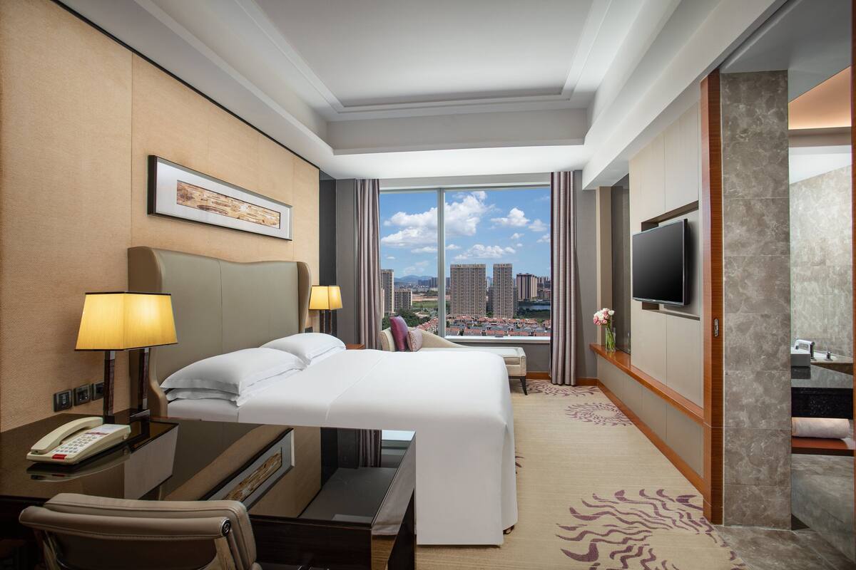 A hotel room with a large bed, a desk with a telephone, a TV mounted on the wall, and a view of the cityscape through large windows.