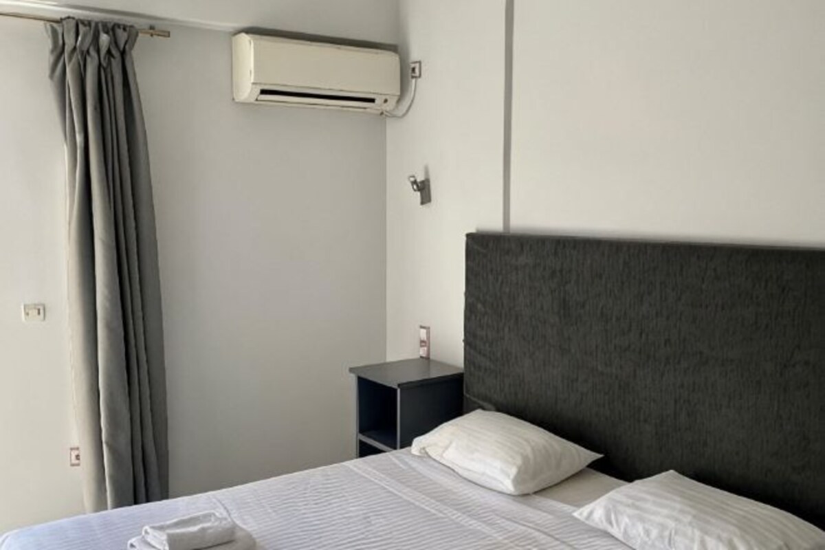 Ett hotellrum med en säng, ett nattduksbord, en väggmonterad luftkonditioneringsanläggning och en gardin.