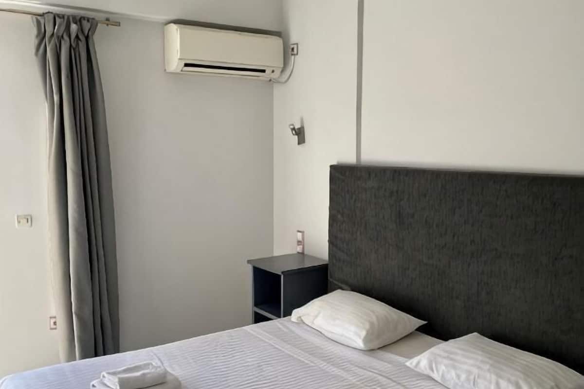 A hotel room with a bed, a nightstand, a wall-mounted air conditioner, and a curtain.