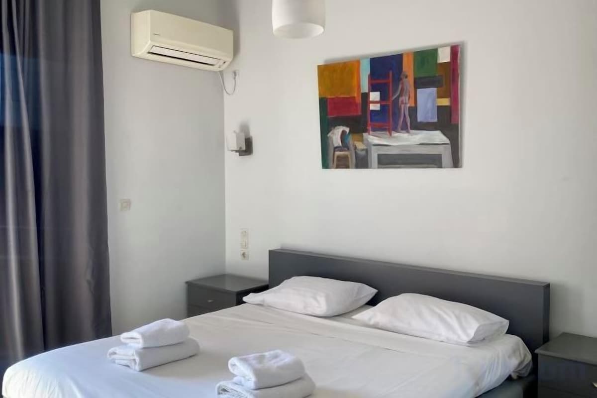 A hotel room with a bed, a bedside table, a wall-mounted air conditioner, and a painting.