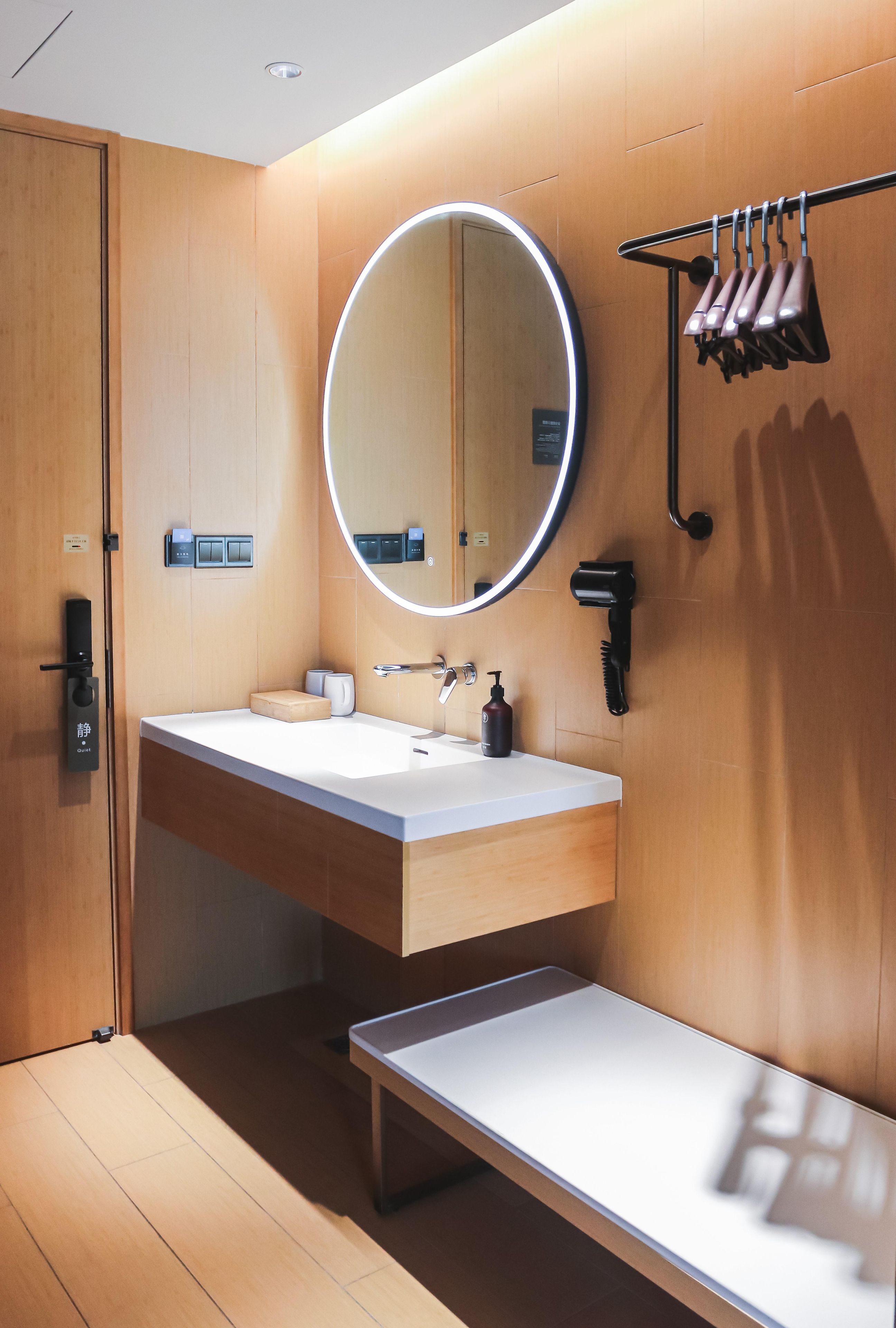 Un baño moderno con tocador de madera, un espejo redondo con iluminación, un secador de pelo y un perchero para toallas.