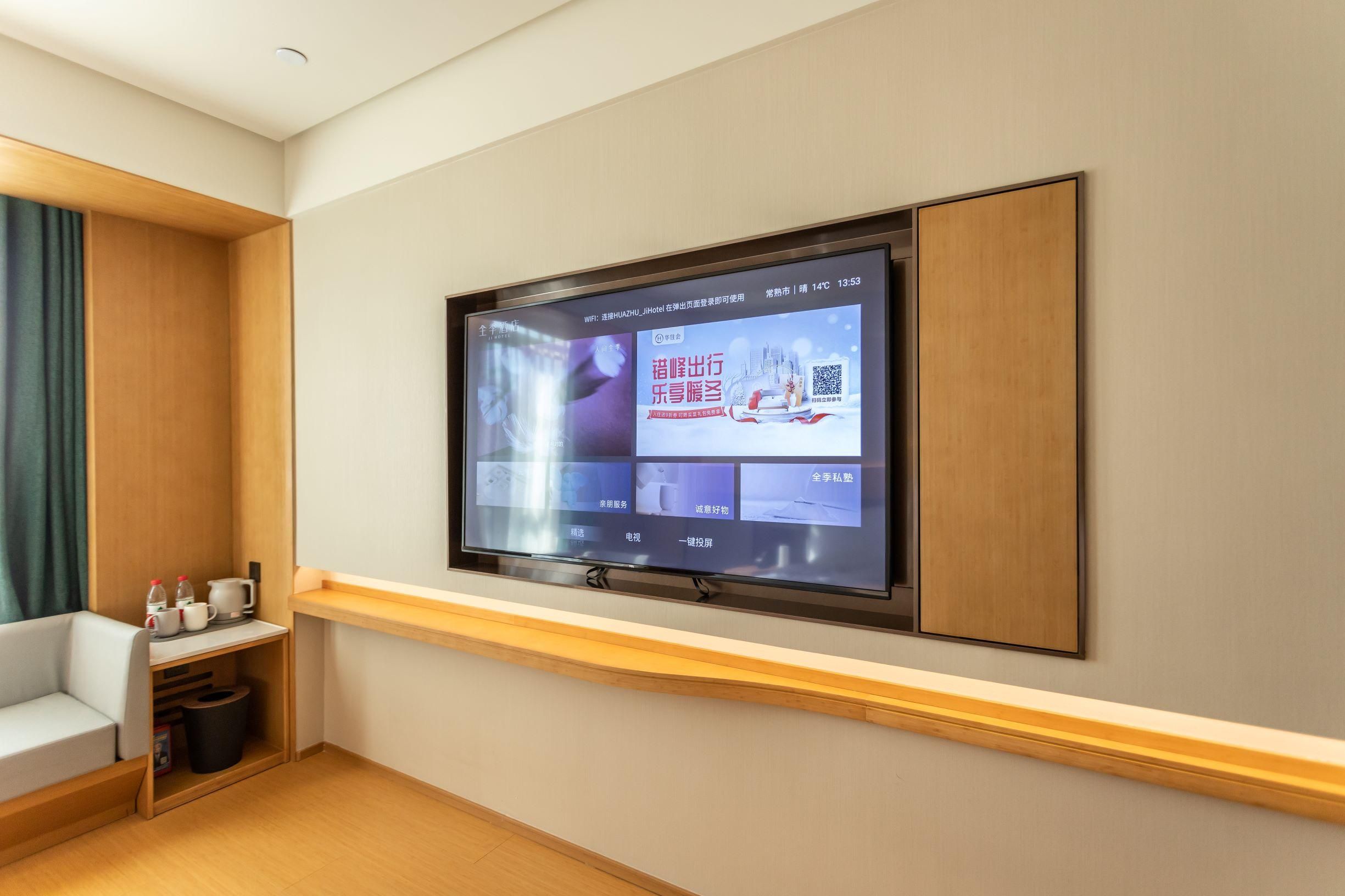 A hotel room with a flat-screen TV mounted on the wall, a wooden bench, a small table with a kettle and cups, and a trash bin.