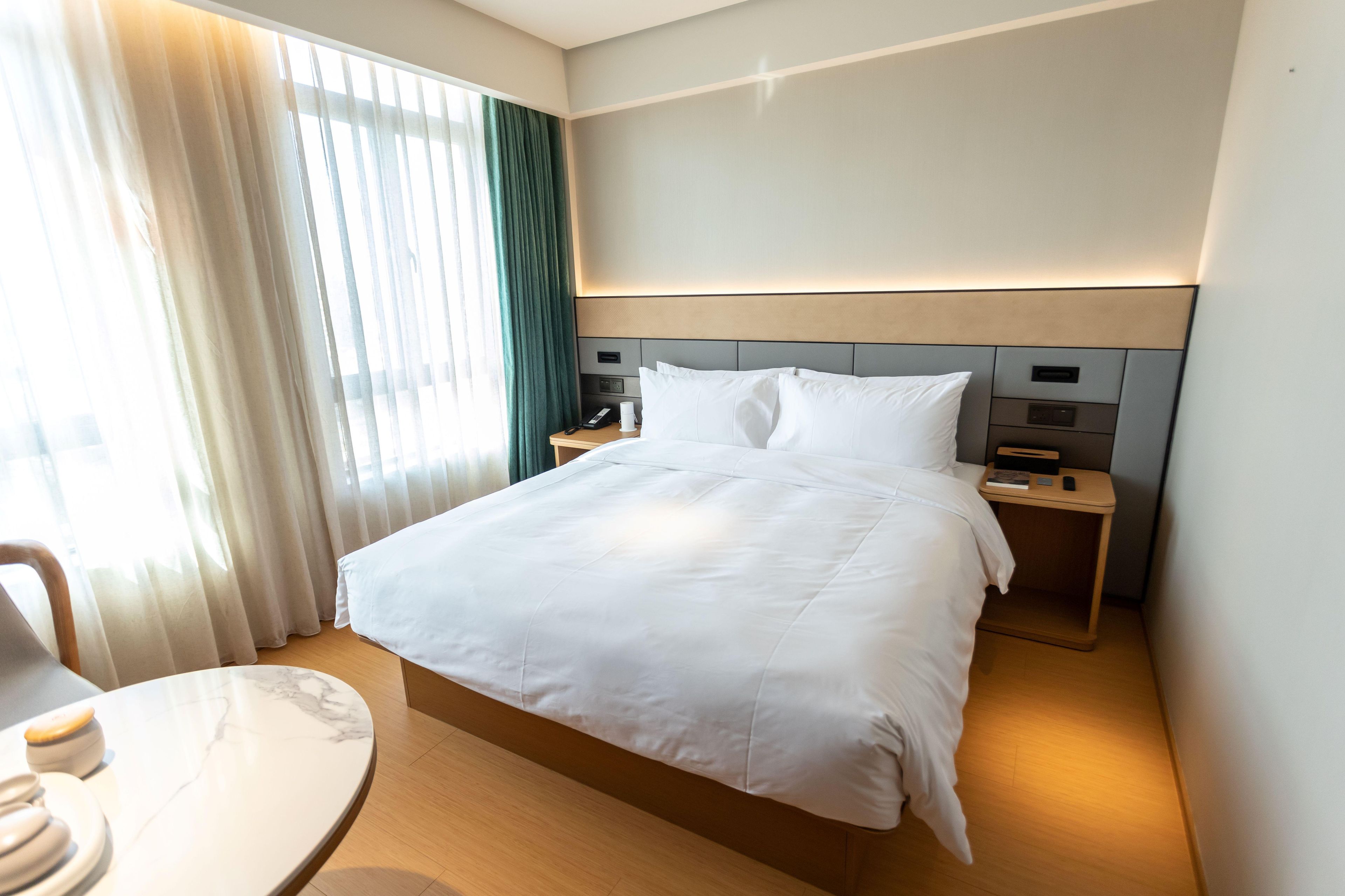 A hotel room with a large bed, a bedside table with a phone and a remote, a small table with a chair, and a window with curtains.