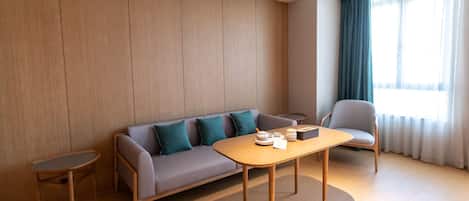Una habitación de hotel moderna con sofá, una mesita, una silla y una ventana con cortinas.
