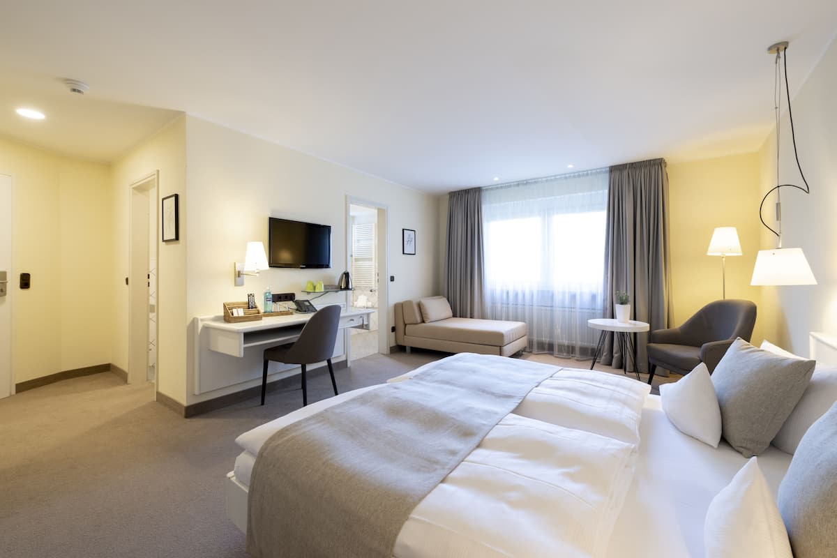 A hotel room with a large bed, a desk with a chair, a TV, and a small table with a lamp.