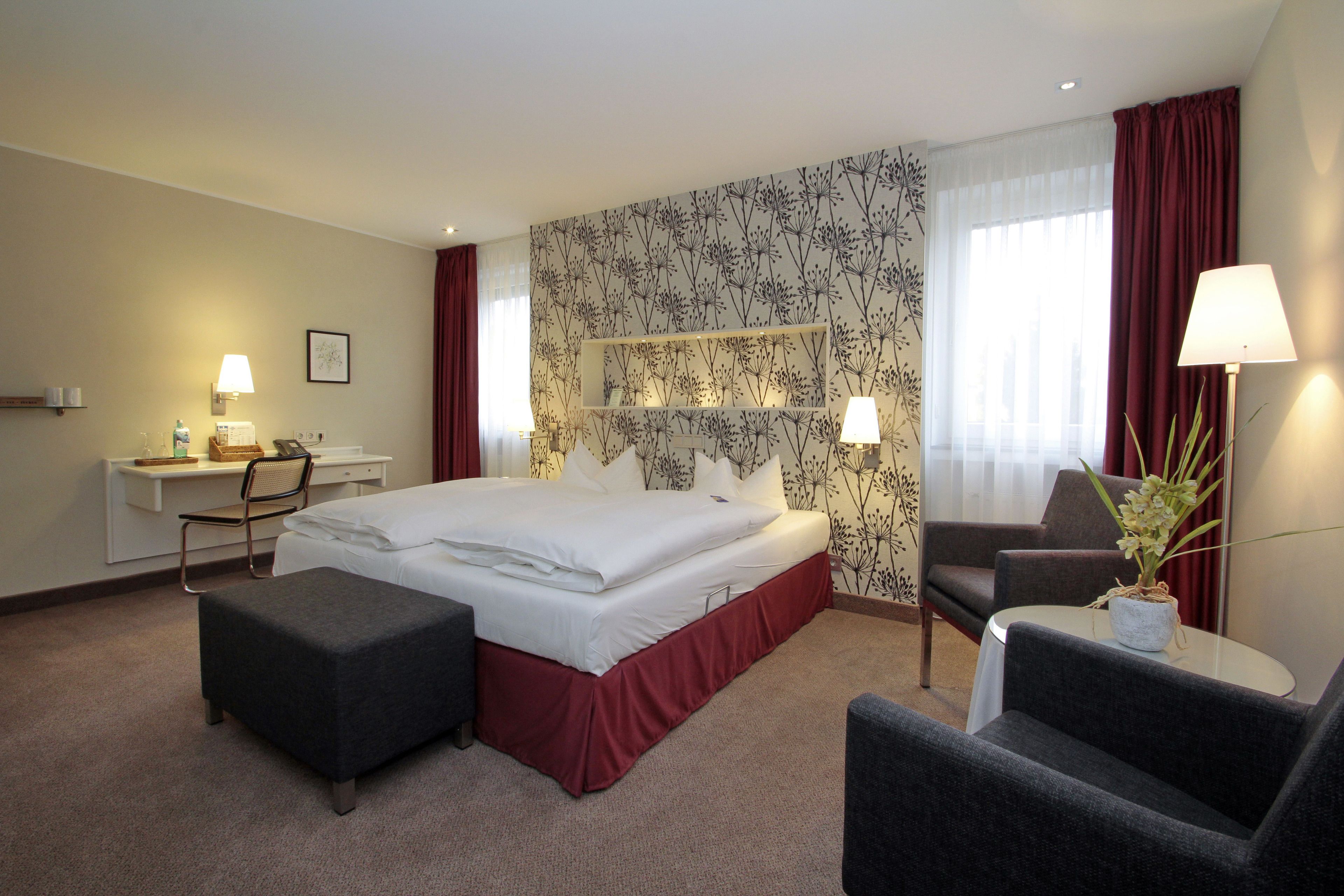 Een hotelkamer met een bed, een bureau, een stoel, een voetenbankje en een klein tafeltje met een lamp.