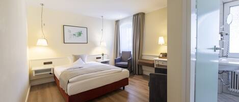 A hotel room with a bed, bedside tables, a chair, a desk, and a bathroom with a sink and mirror.