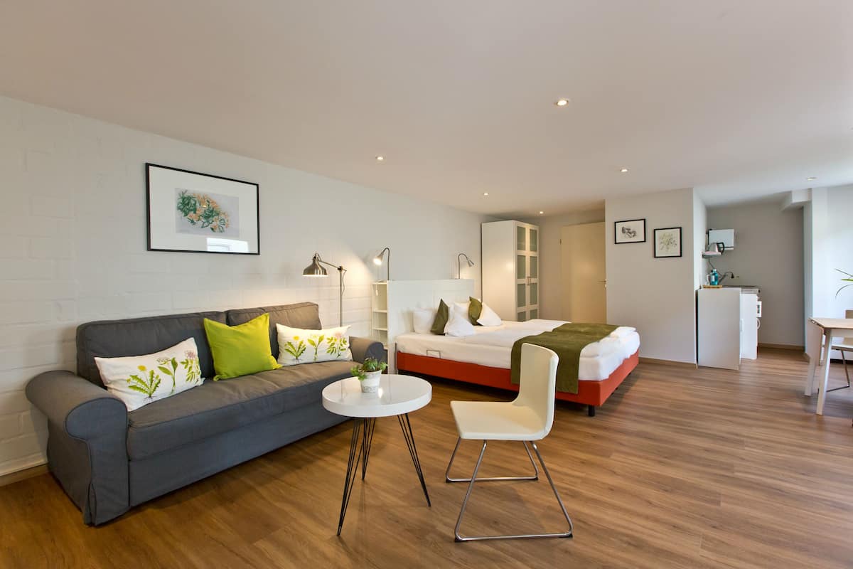 A modern hotel room with a grey sofa, a white chair, a small round table, a bed with a green blanket, and a white wardrobe.