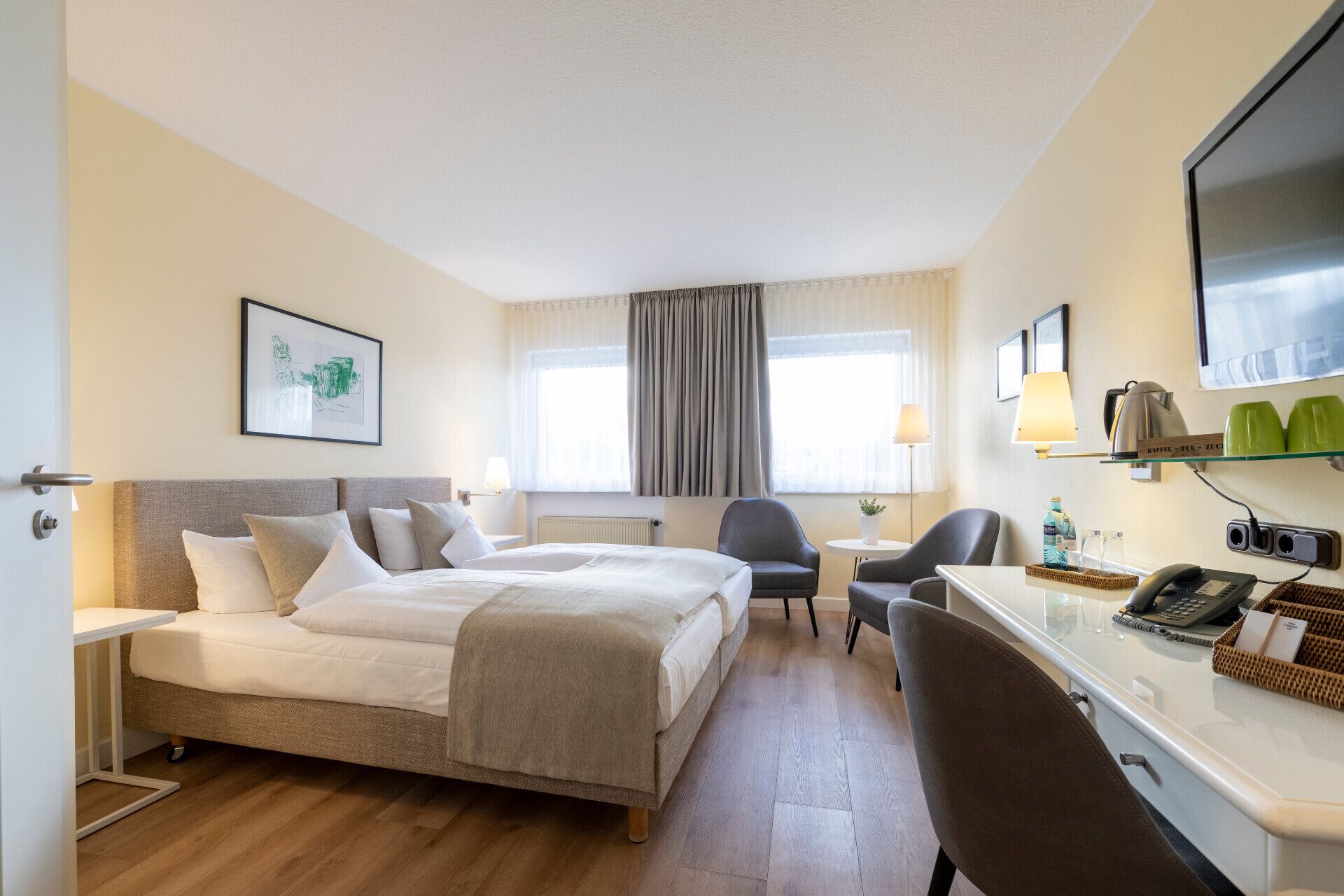 A hotel room with a bed, two chairs, a desk with a telephone and a lamp, and a television.