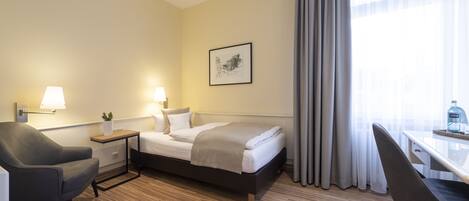 Una camera d'albergo con un letto, una scrivania, una sedia, un comodino con una lampada e una finestra con tende.