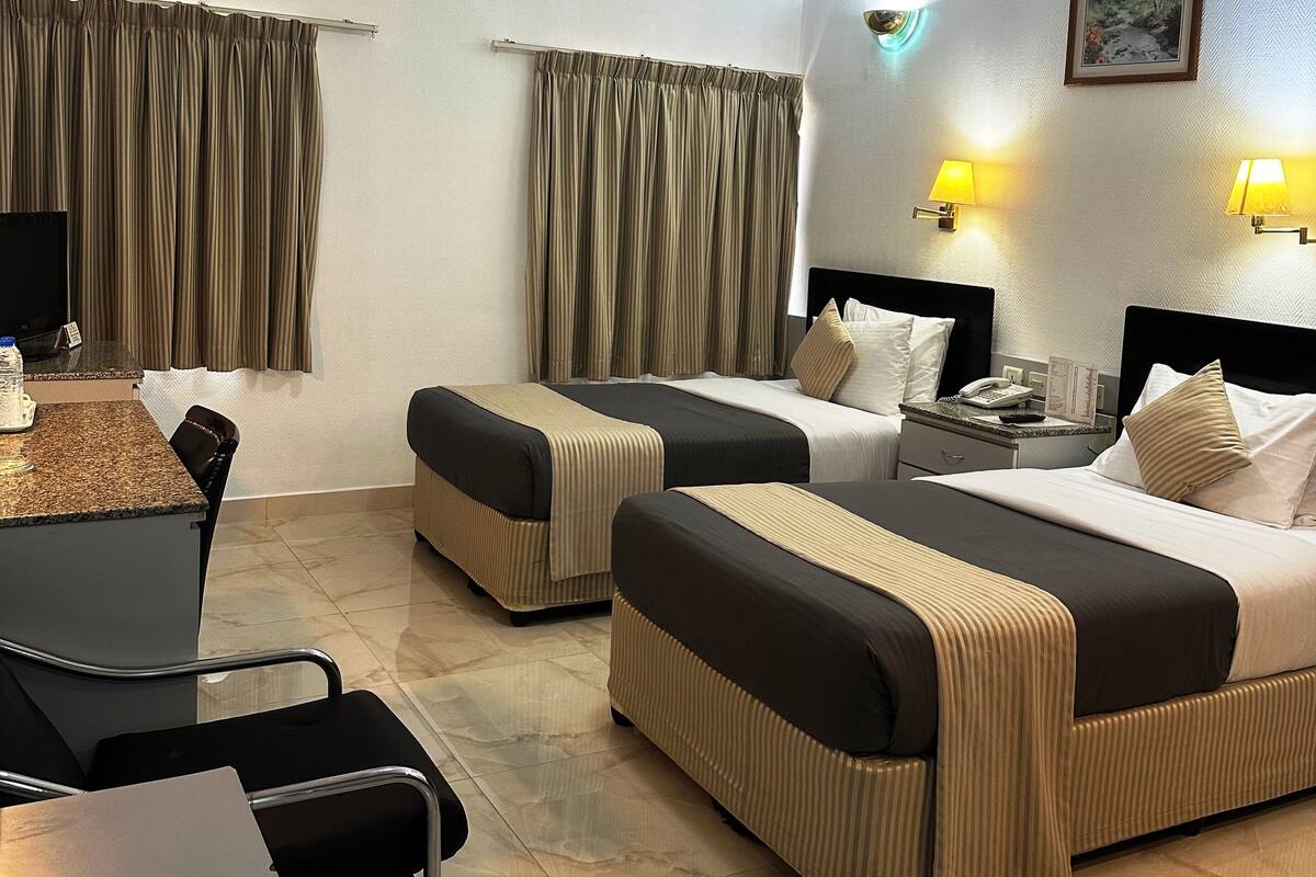 A hotel room with two beds, a desk, a chair, a table, a telephone, and two wall-mounted lamps.