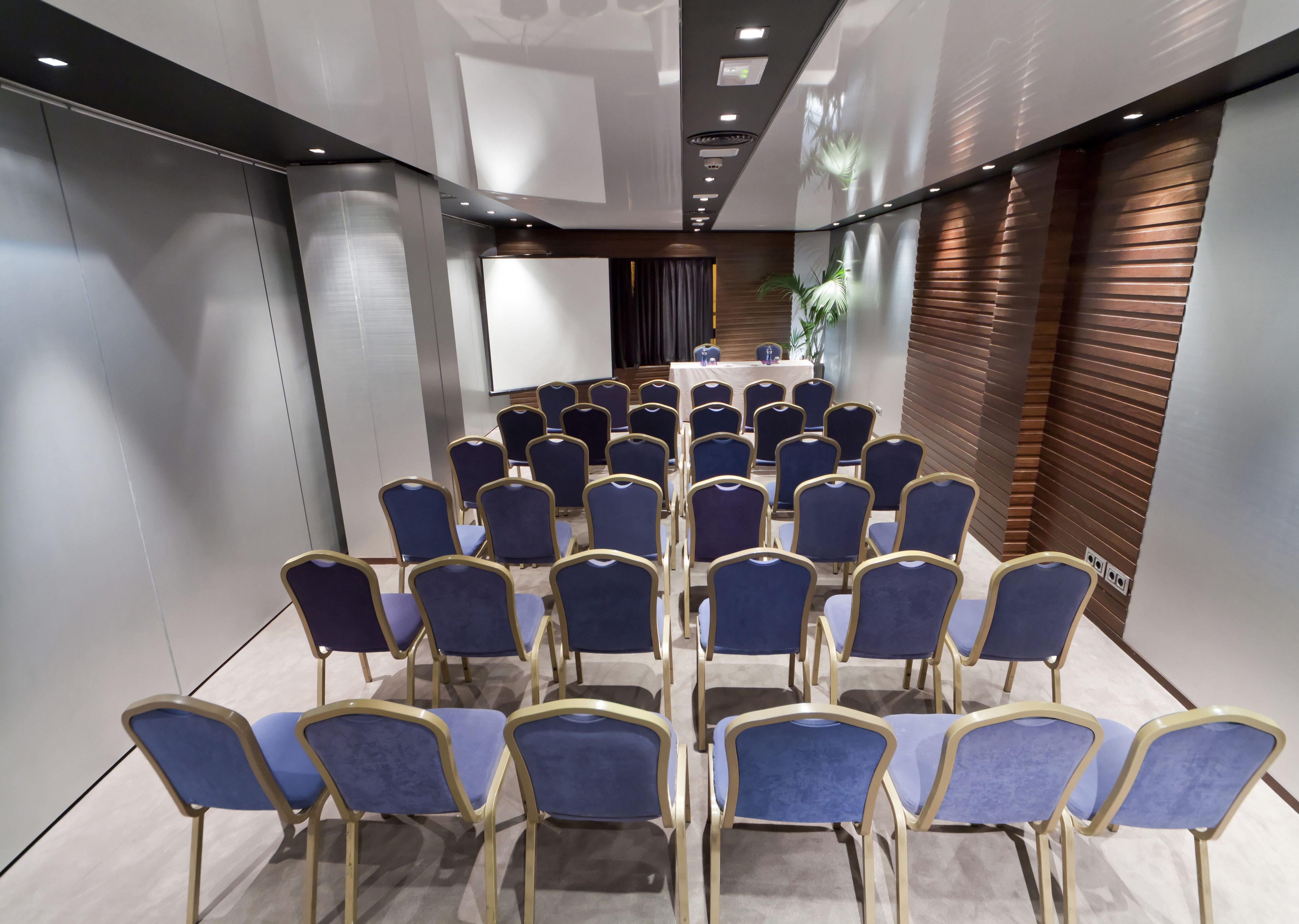 A conference room with rows of blue chairs and a white table at the front.