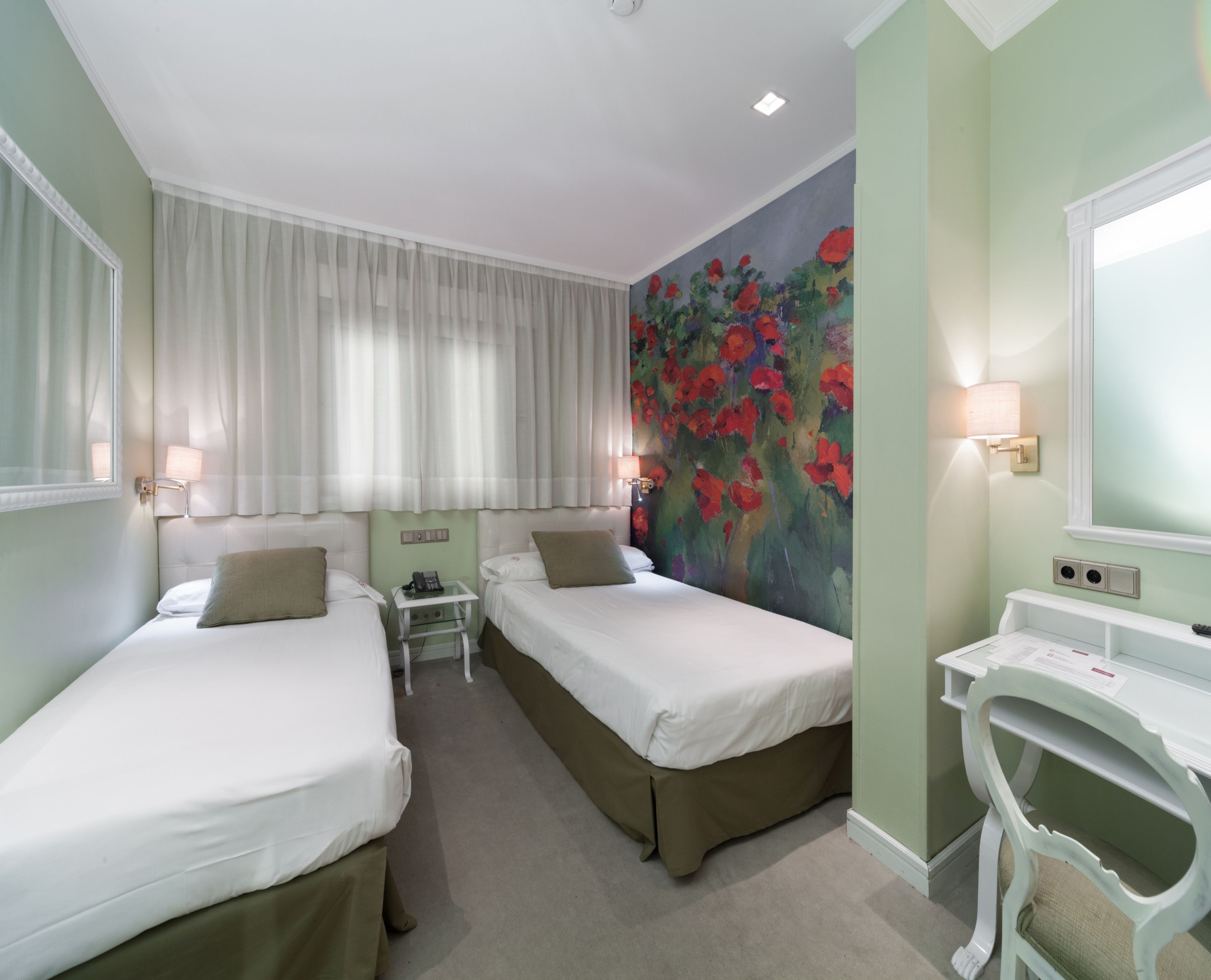 Habitación de hotel con dos camas, una mesita de noche con lámpara, un cuadro de flores rojas en la pared y un escritorio blanco con espejo.