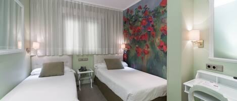 A hotel room with two beds, a small bedside table with a lamp, a painting of red flowers on the wall, and a white desk with a mirror.