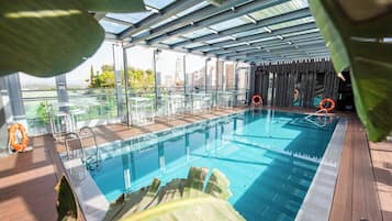 An indoor swimming pool with clear glass walls, a wooden deck, and a view of the cityscape.