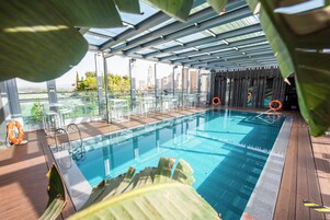 En indendørs swimmingpool med glasvægge, en træterrasse og udsigt over bybilledet.