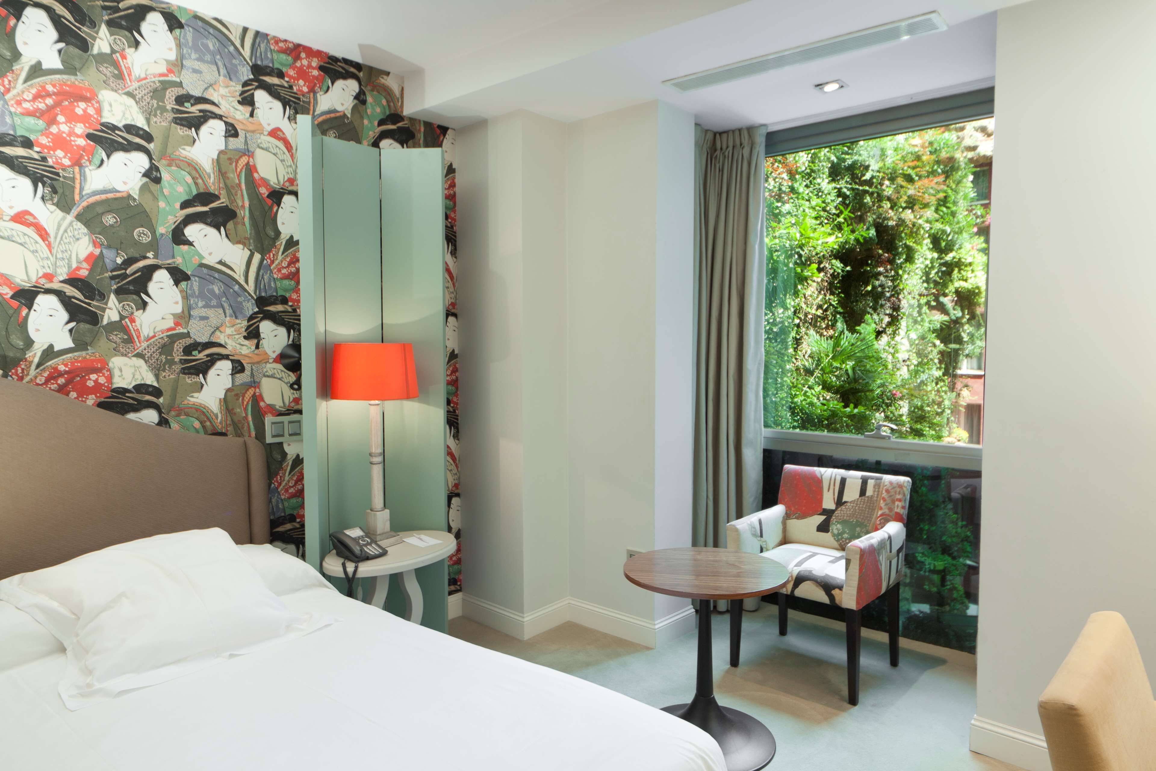 Ein Hotelzimmer mit Bett, Schreibtisch, Stuhl, Lampe und einem großen Fenster mit Blick auf Grünflächen.