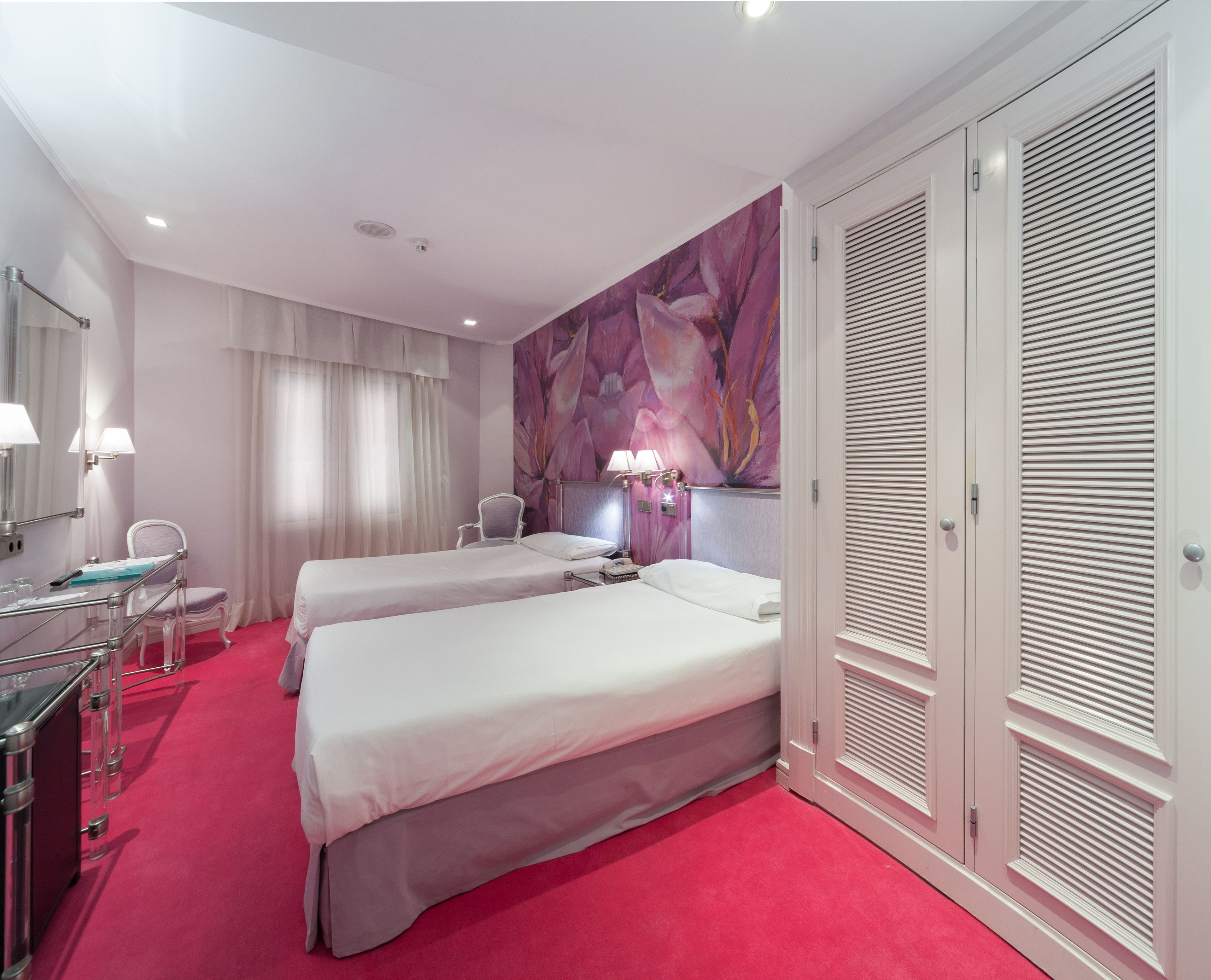 A hotel room with two beds, a vanity with a mirror, a chair, a desk, and a large window with blinds.
