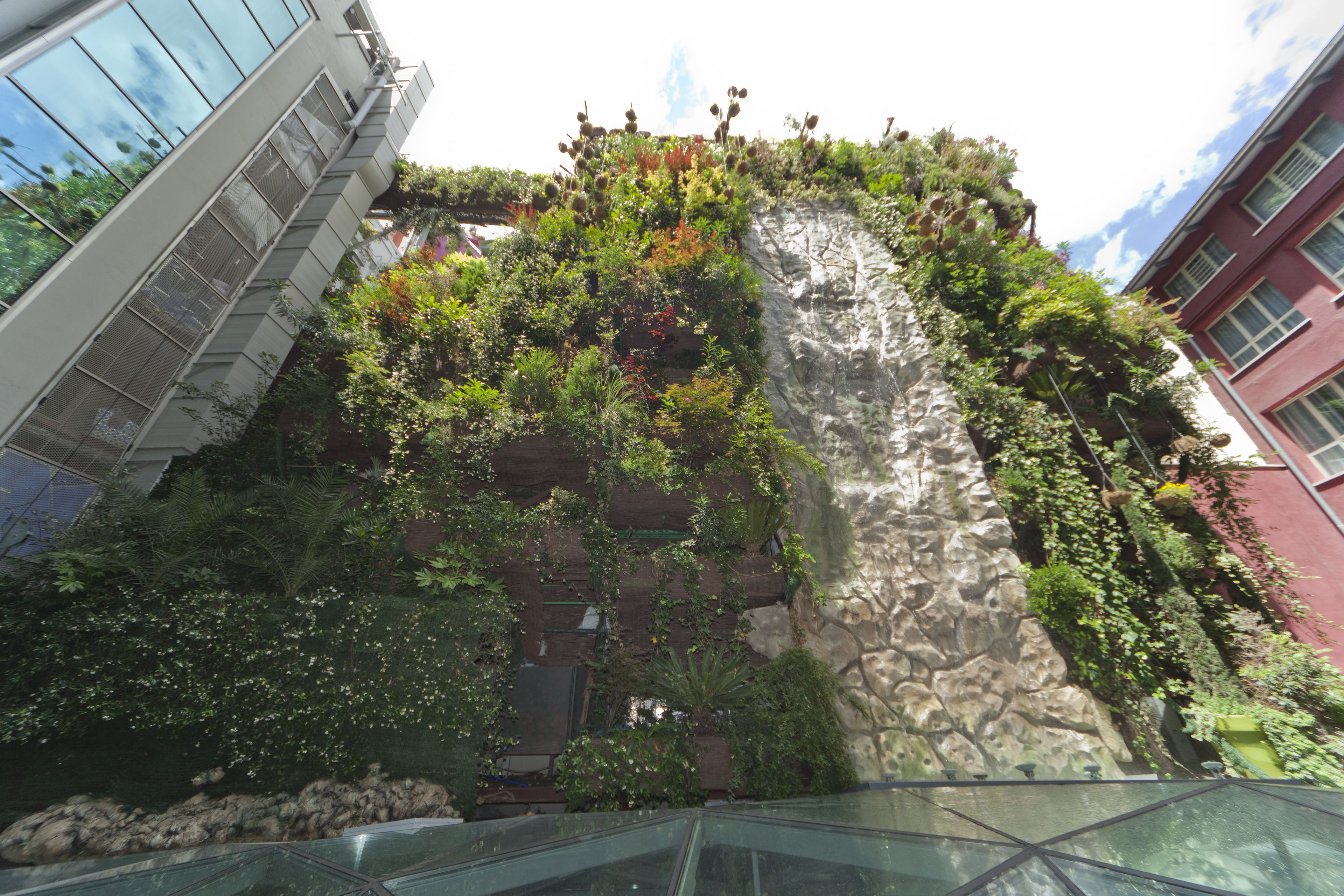 A building with a rock facade covered in greenery, modern glass structures, and a red building in the background.