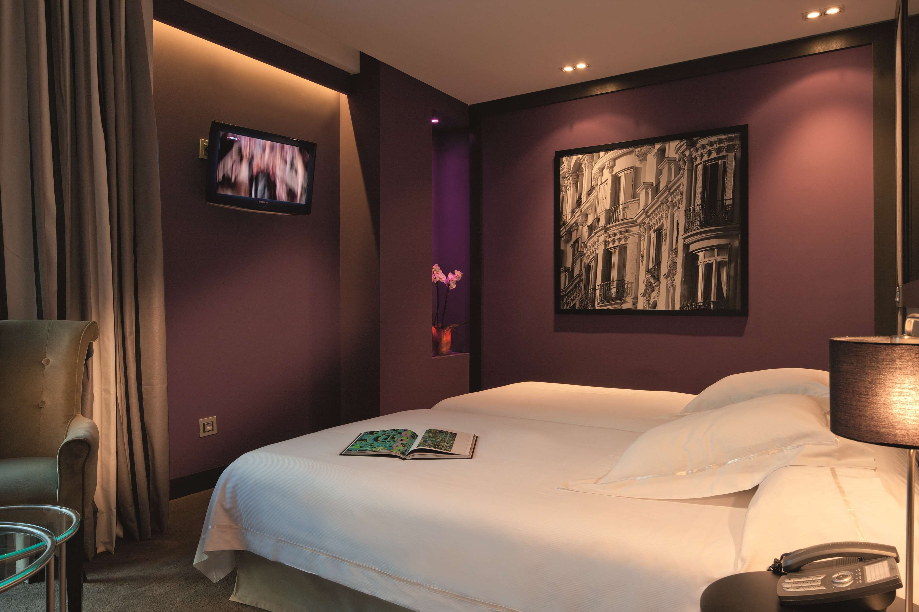 A hotel room with a bed, a chair, a table, a TV, and a painting on the wall.