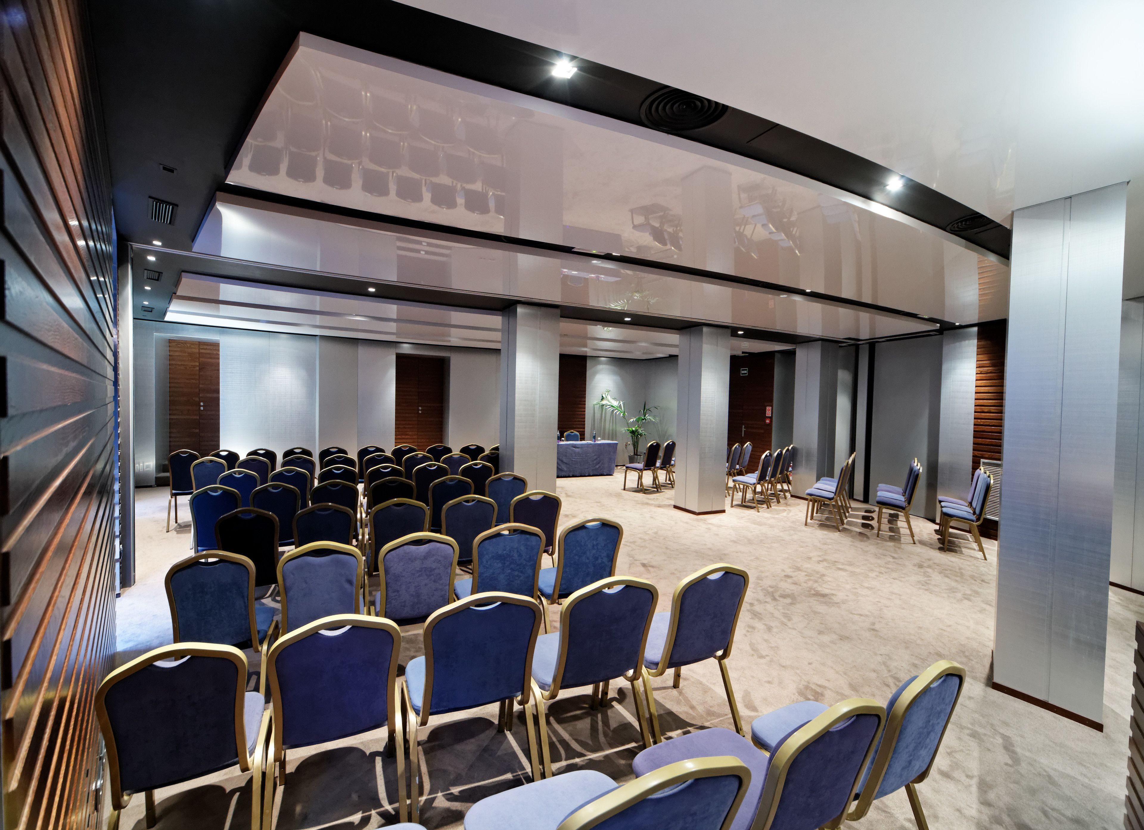 Un salón de conferencias con filas de sillas azules, un escenario y un ventanal grande.