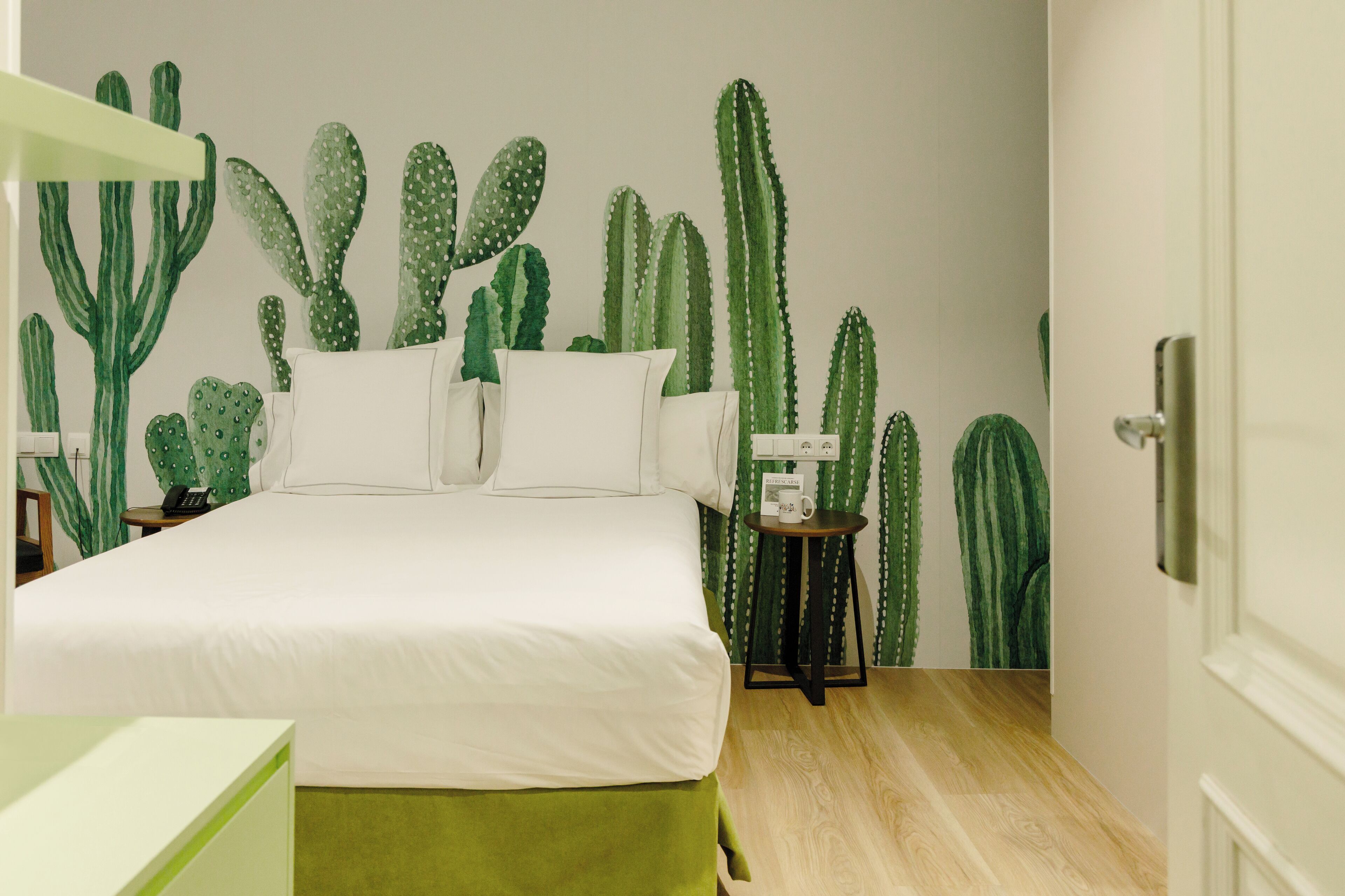 A bedroom with a bed, pillows, a nightstand, a small table with a lamp, and a wall with cactus artwork.