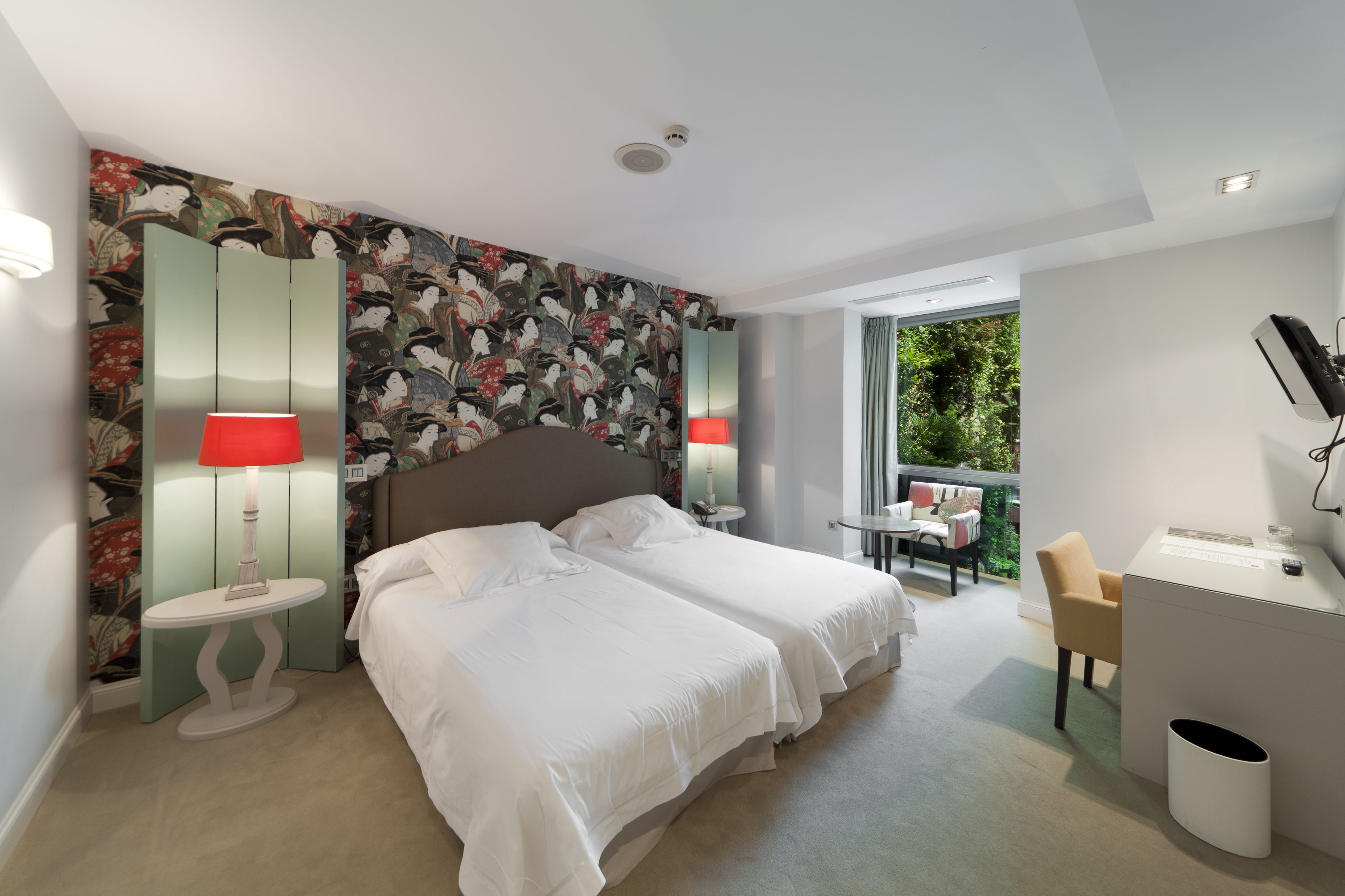A hotel room with two beds, a desk, a chair, and a view of greenery outside.
