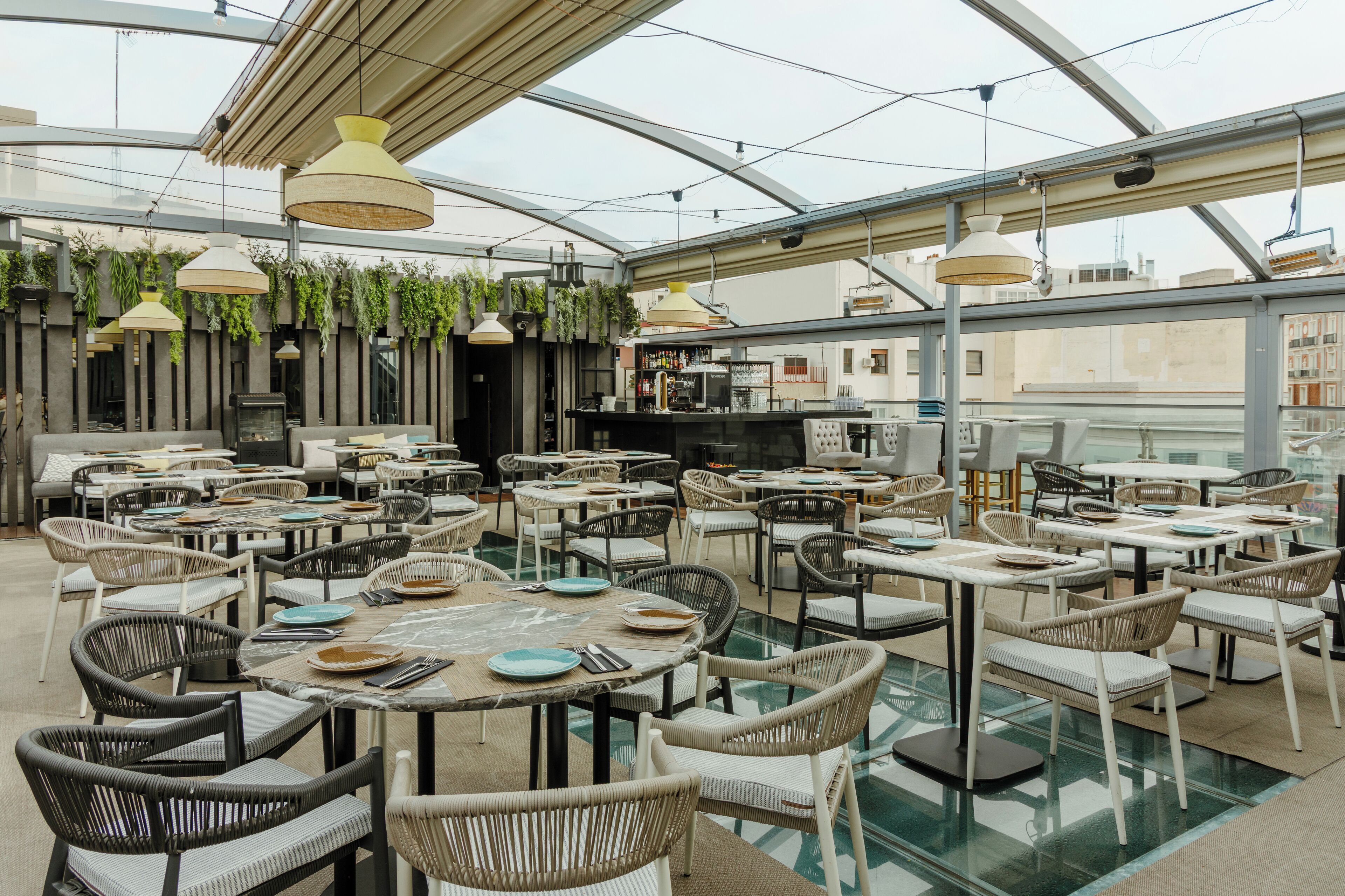 A rooftop restaurant with multiple tables and chairs, a bar area, and hanging plants.