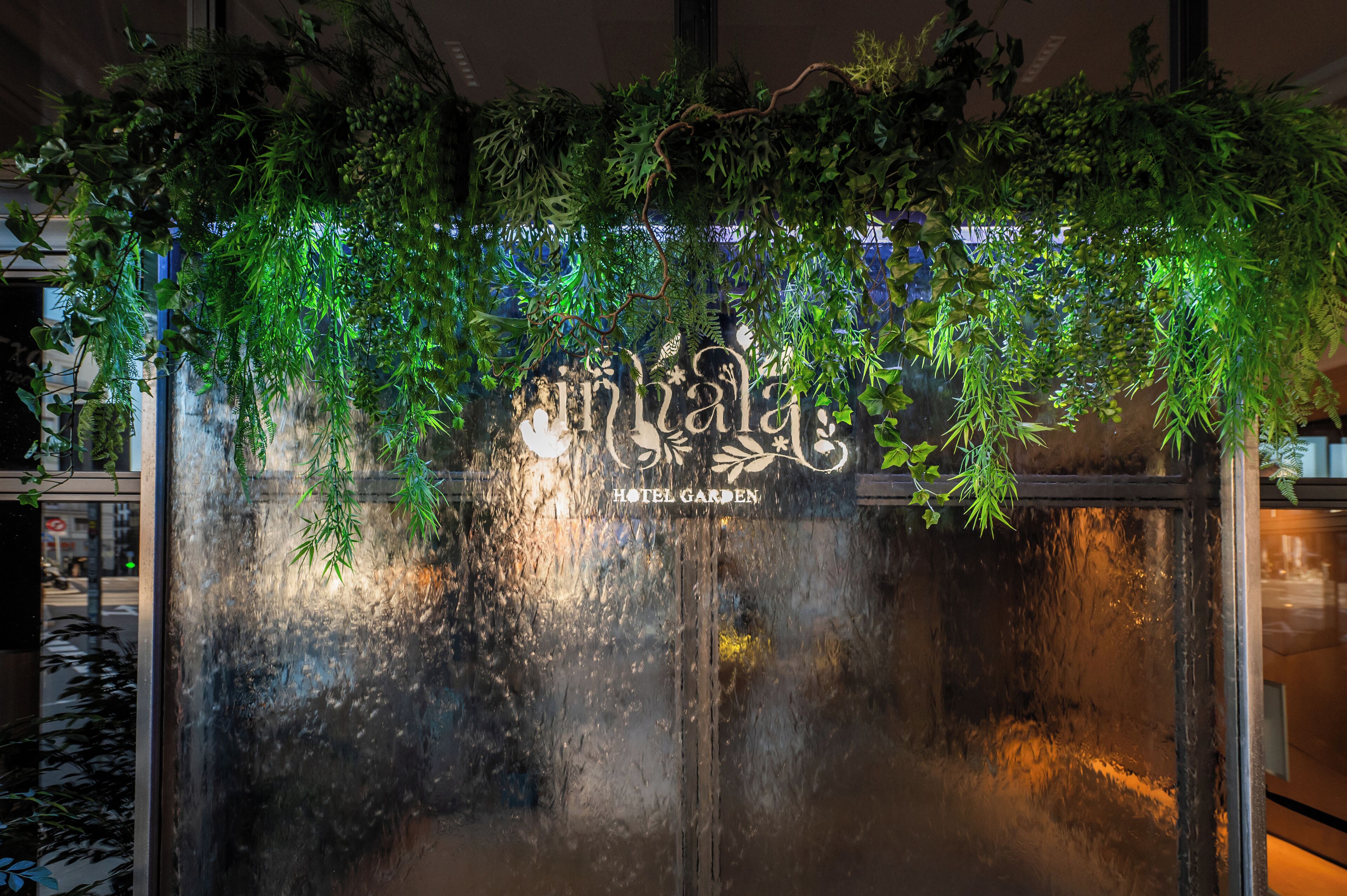 L’entrée d’un hôtel, ornée de plantes suspendues, est surmontée d’une enseigne indiquant « Hôtel Garden ».