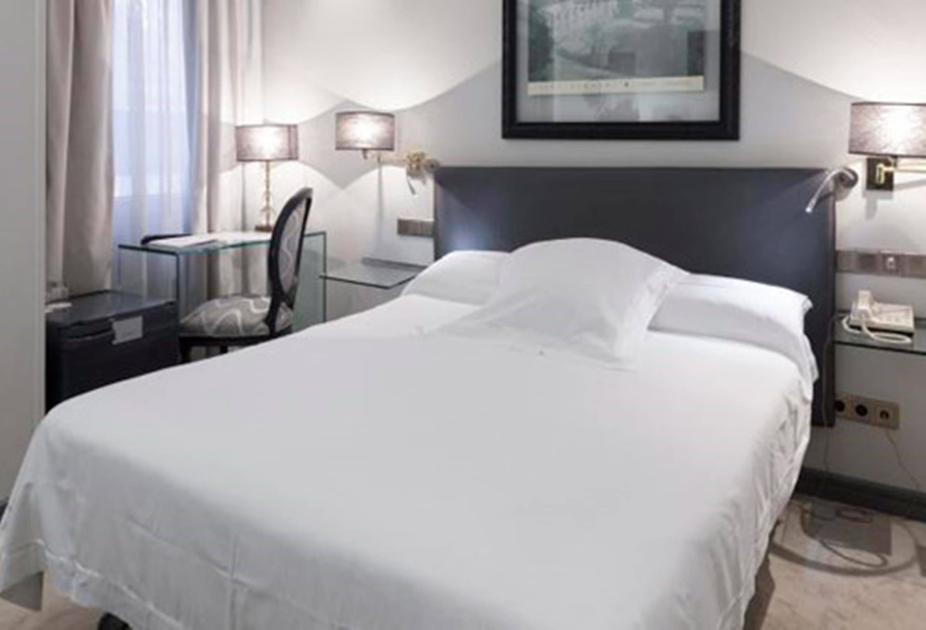 Habitación de hotel con una cama bien tendida, un escritorio con una silla, una lámpara y un cuadro en la pared.