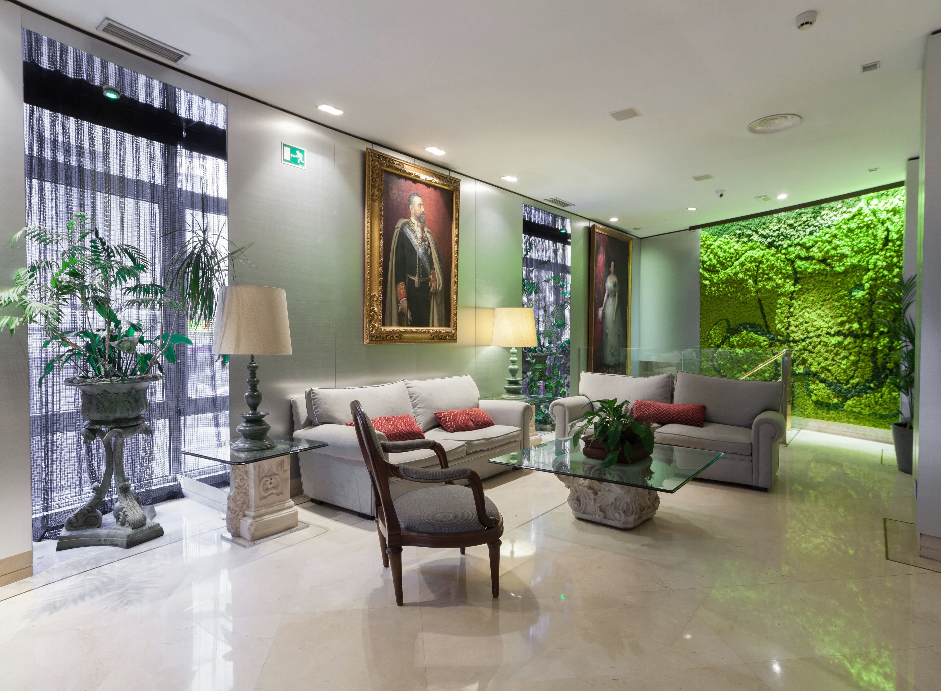 A modern lobby with two sofas, a glass coffee table, and a decorative chair. There are potted plants and a large framed portrait on the wall.