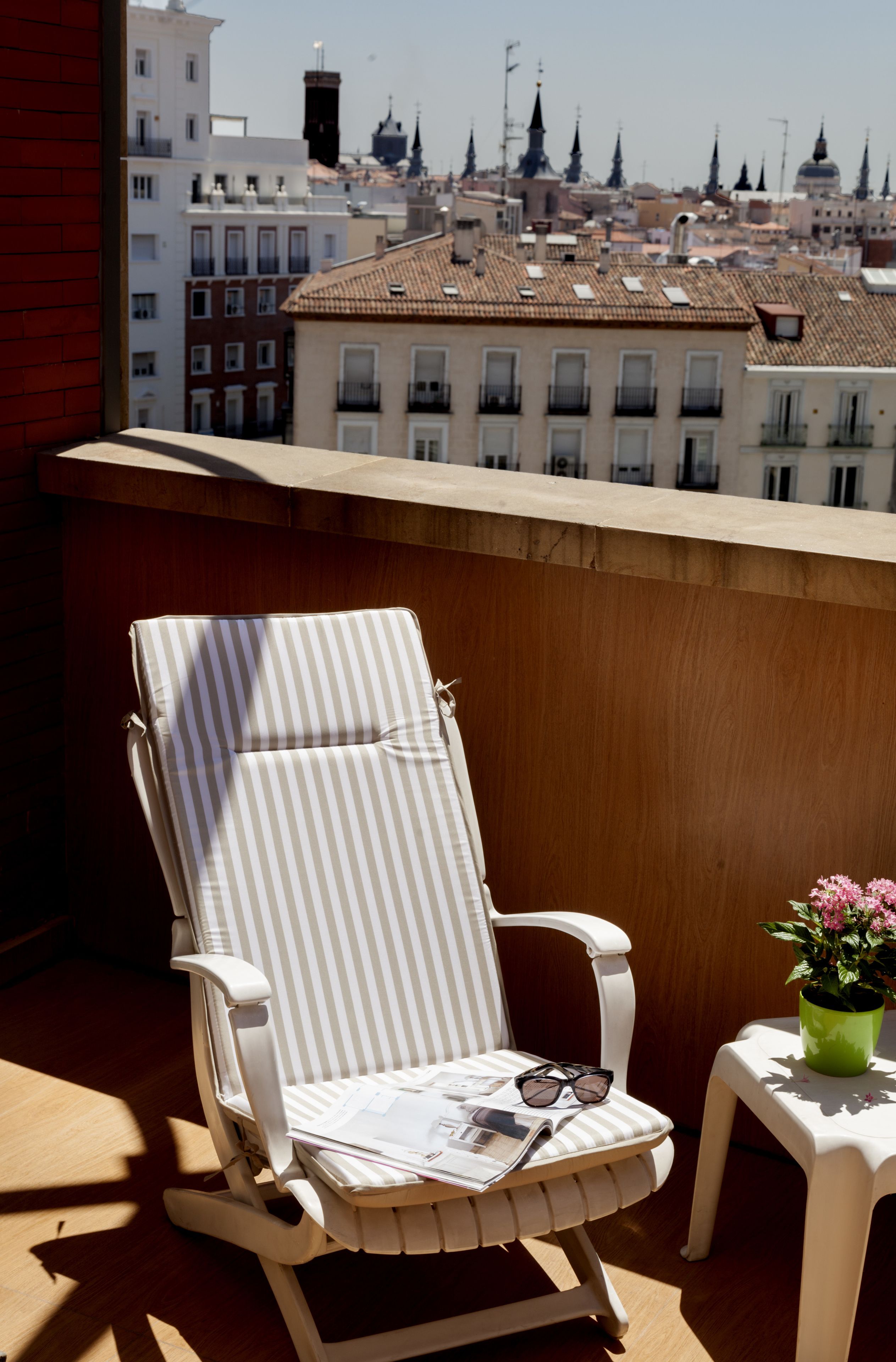 A balcony with a striped lounge chair, a small table with a potted plant, and a cityscape view in the background.