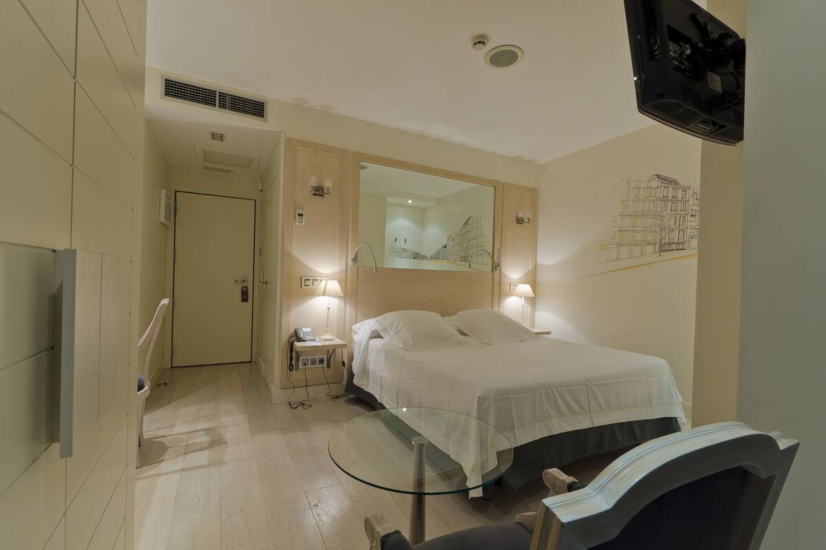 A hotel room with a bed, bedside lamps, a glass table, a chair, a TV, and a door leading to another room.