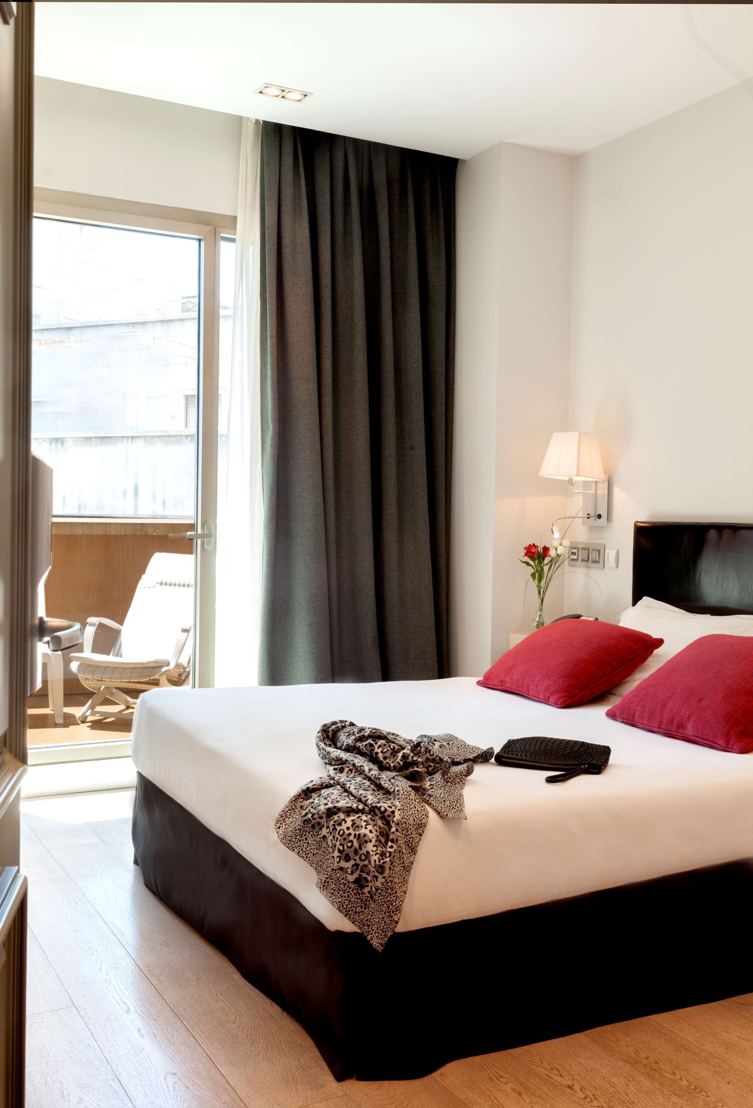 A hotel room with a large bed, a chair, a nightstand with a lamp, and a view of the outside.