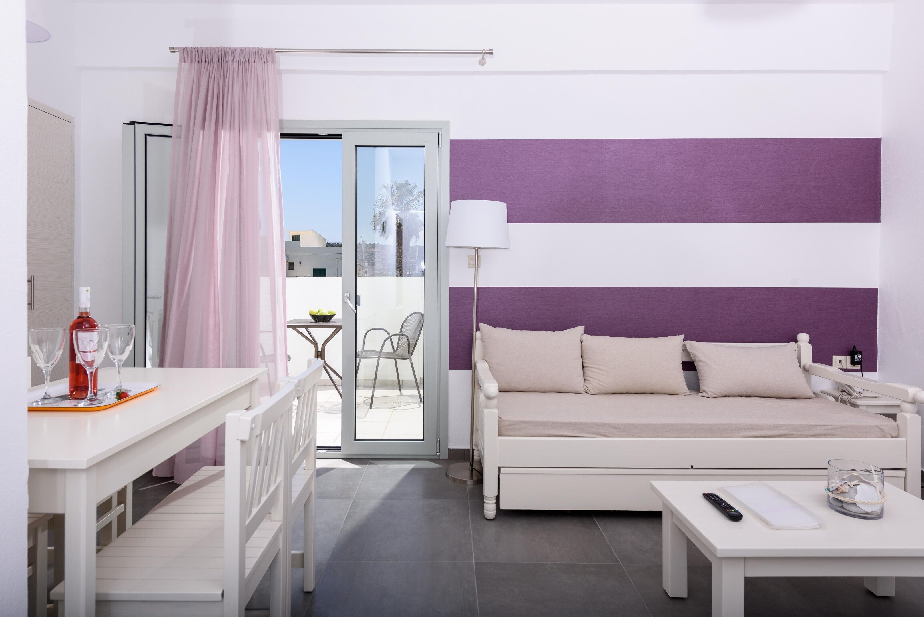 A modern hotel room with a white bed, a white desk with a bottle and glasses, a purple-striped wall, and a sliding glass door to the balcony.