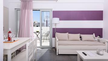A modern hotel room with a white bed, a white desk with a bottle and glasses, a purple-striped wall, and a sliding glass door to the balcony.