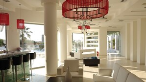A spacious living area with a bar, white sofas, and a large window overlooking a pool and palm trees. - Angela Studios & Apartments (Agios Nikolaos)